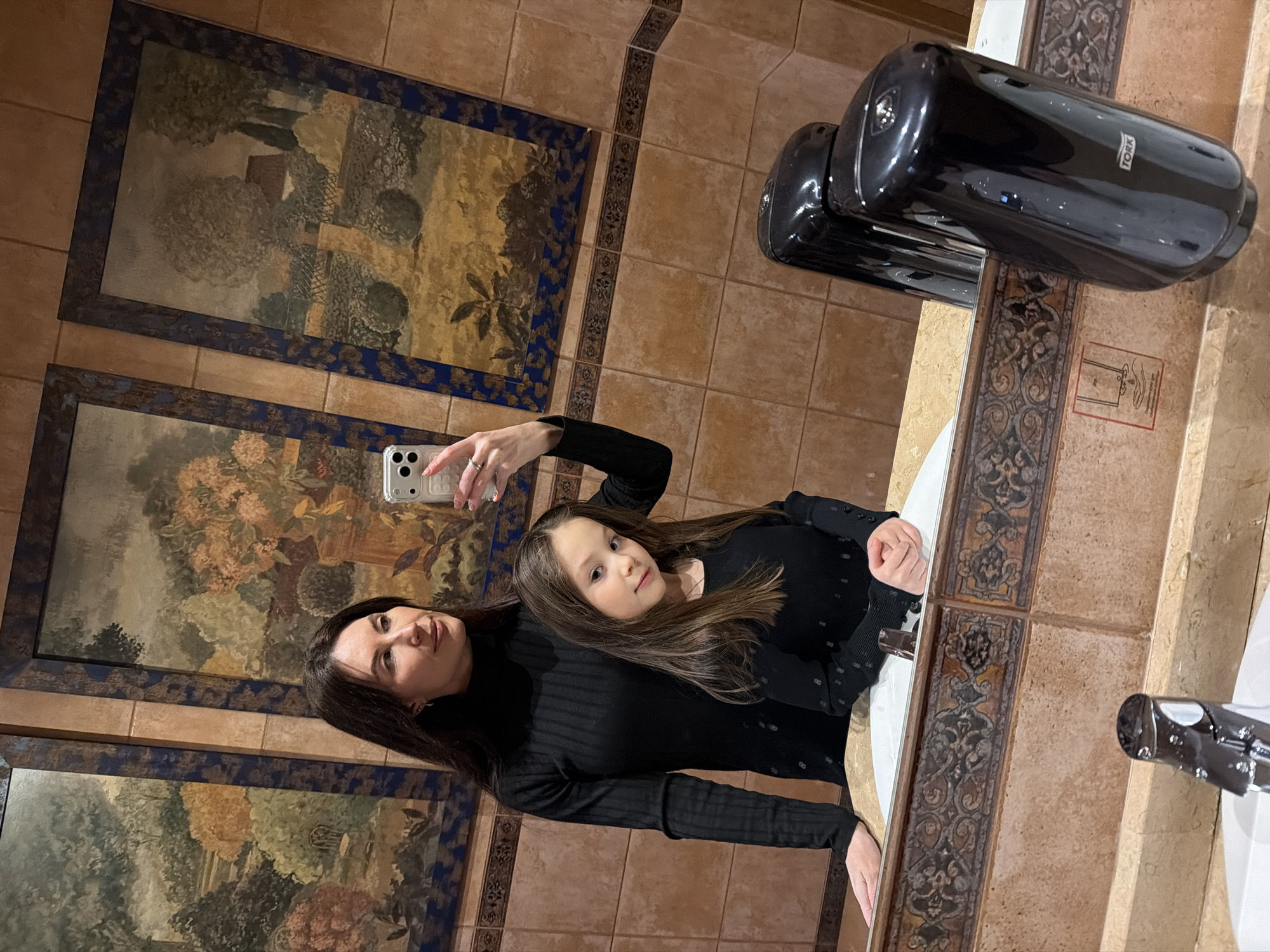 A woman and a child are taking a selfie in front of a mirror in a bathroom. The bathroom has ornate tile work and framed landscape art on the walls. A black Tork hand dryer is mounted on the wall near the mirror. The photograph was taken in Perm, Russia.