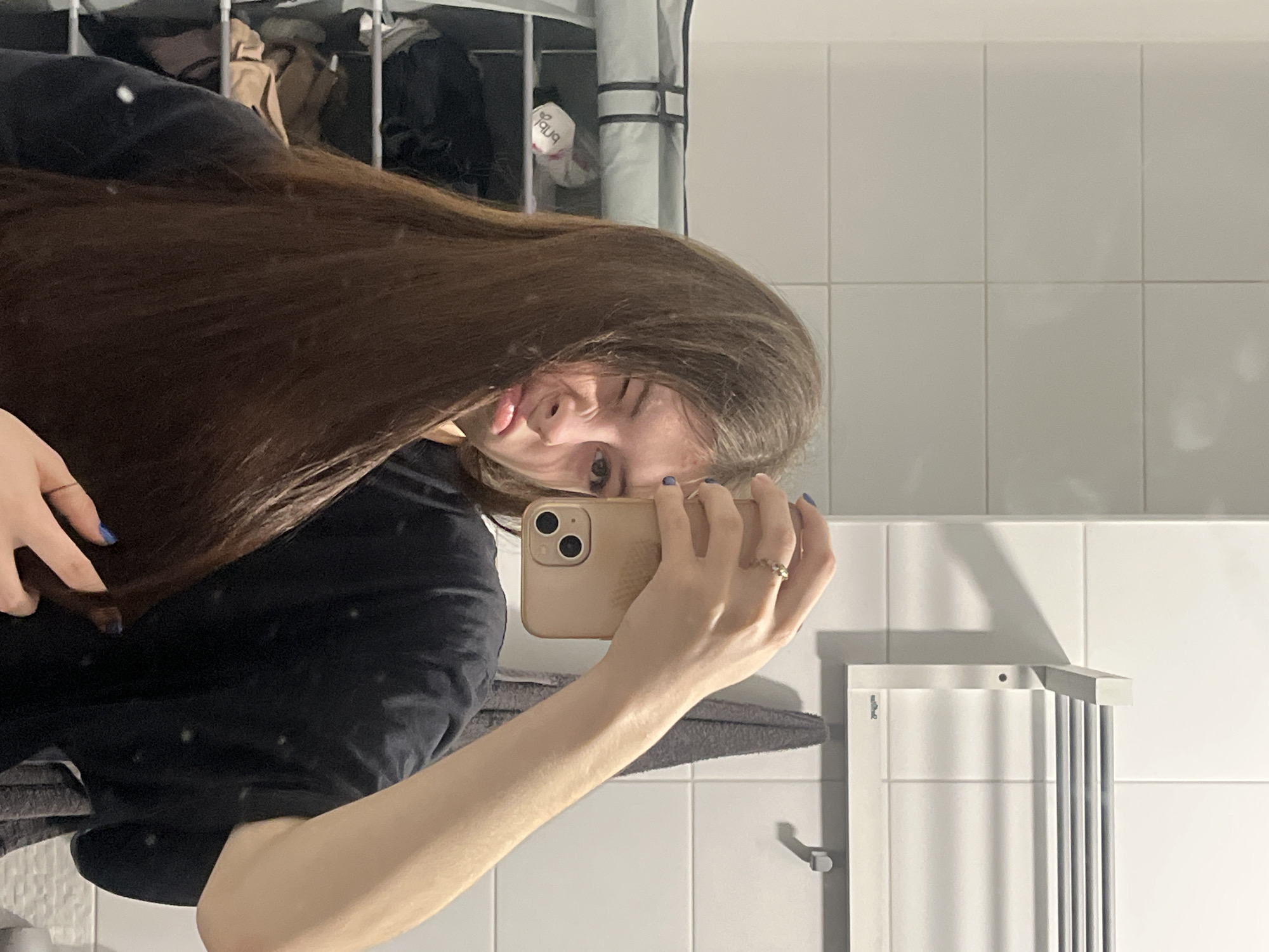 A woman is taking a selfie in what appears to be a bathroom. She is holding a tan iPhone in her right hand. Her hair is long and brown. She is wearing a black t-shirt. The bathroom has white tiled walls and a towel rack. A storage shelf with items inside is visible in the background. The image does not contain sufficient information to determine a location in Saint Petersburg, Russia.