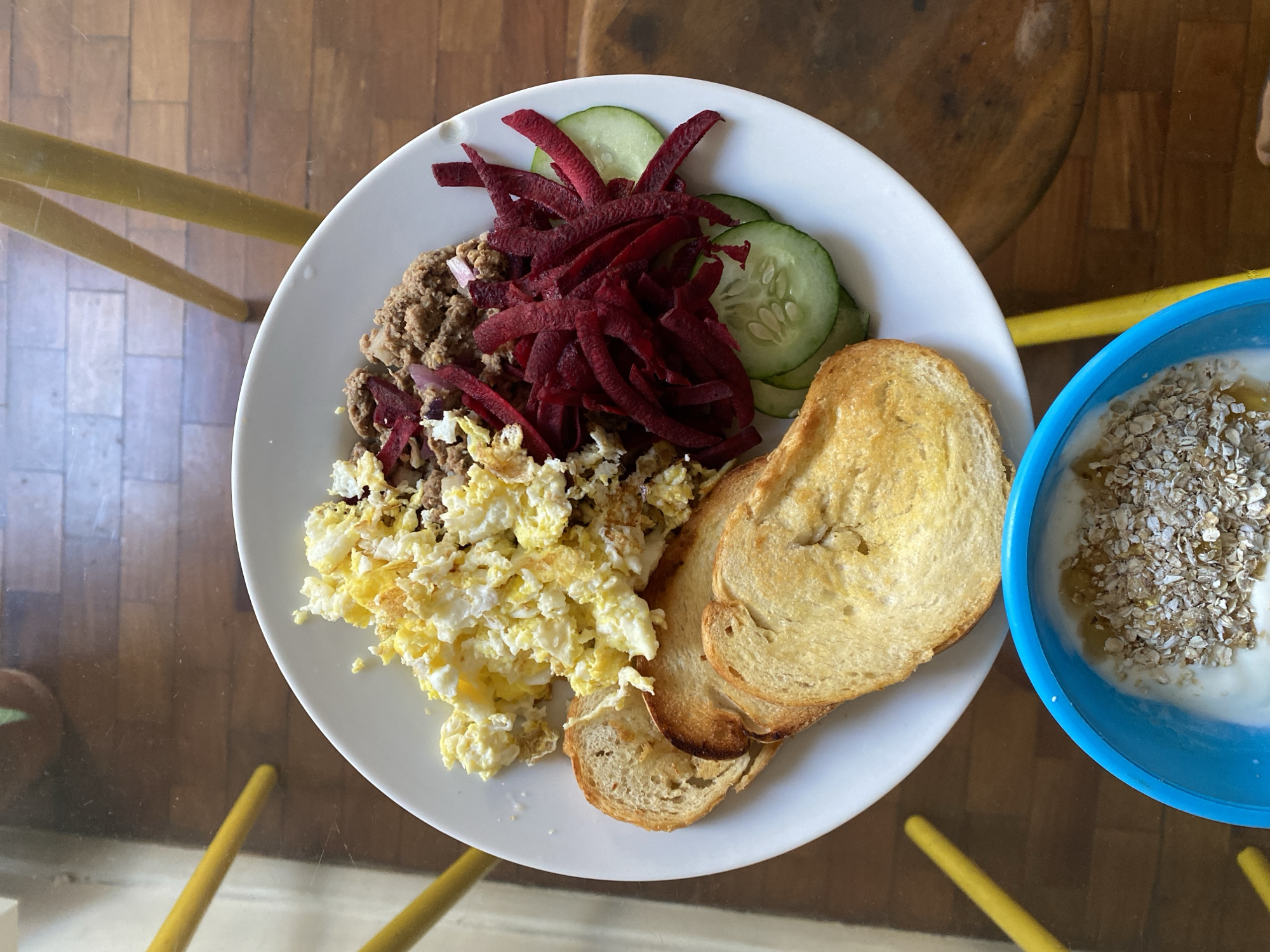 A top-down view shows a plate of food on a glass table, along with a blue bowl. The plate contains scrambled eggs, ground meat, julienned beets, cucumber slices, and toasted bread. The blue bowl appears to hold yogurt with oats. The scene is located in São Paulo, Brazil. The objects are arranged on a table with yellow legs.