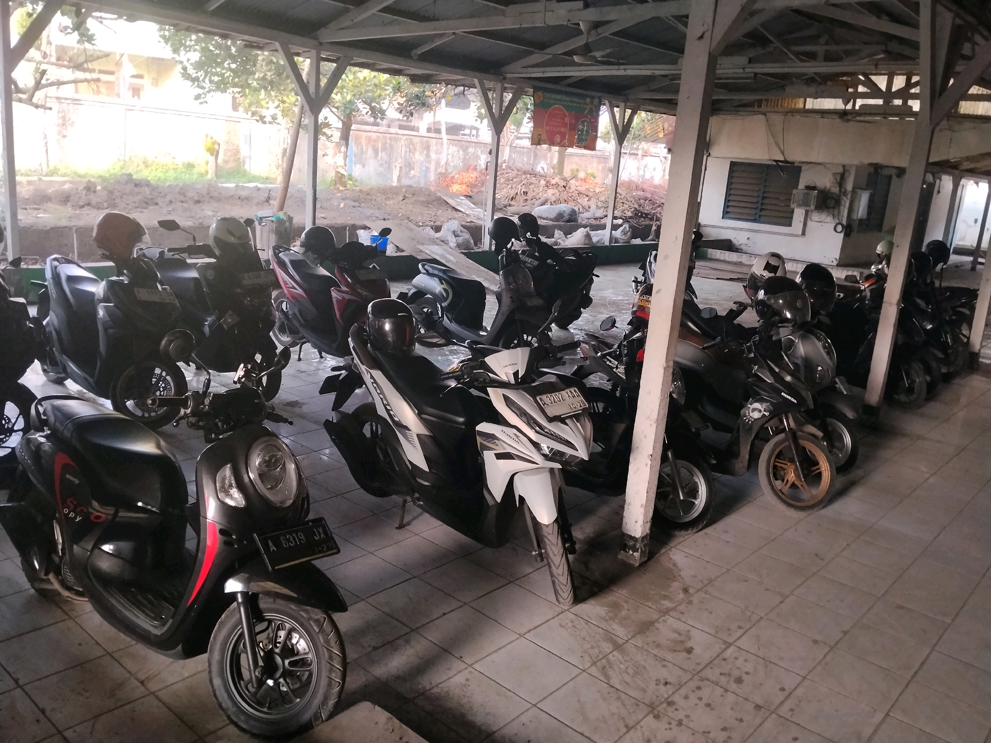The image shows a covered parking area filled with parked motorcycles, located in the city of Suka Nagara, Indonesia. The parking area has a light-colored, tiled floor and is supported by wooden pillars and a corrugated metal roof. The motorcycles are of various models and colors, predominantly black, white, and red, with some helmets placed on them. Some motorcycles have license plates visible, indicating registration. In the background, there is a wall with a window and a sign with text and graphics, suggesting this might be a public or commercial establishment. The outdoor area beyond the covered space appears to be under construction or in a state of disarray, with piles of dirt and debris visible. The lighting suggests it is daytime, with natural light filtering into the parking area. There are no people visible in the image.
