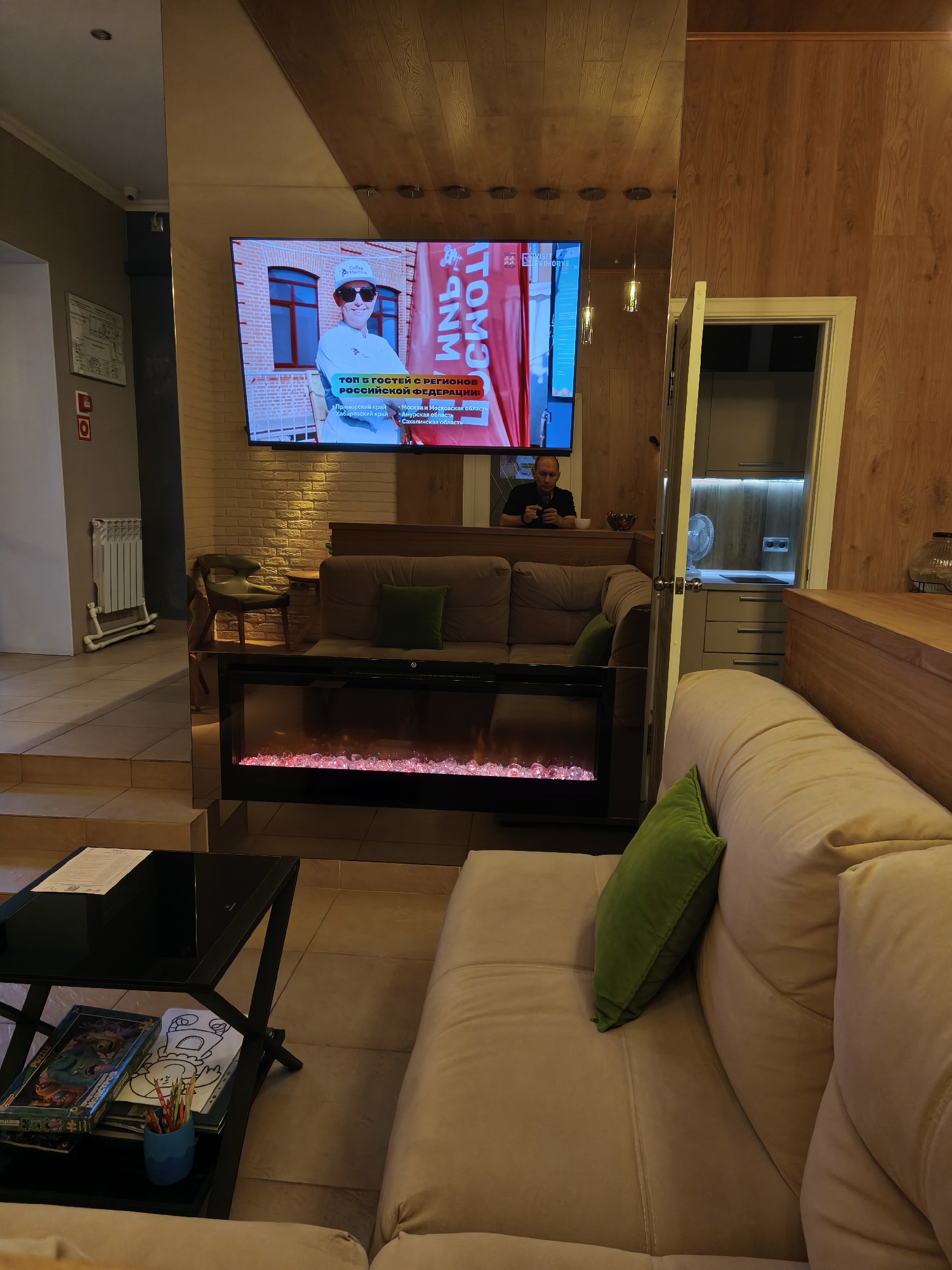 The image depicts the interior of a modern establishment, possibly a cafe or lounge, with an emphasis on comfort and visual appeal. A large flat-screen television mounted on a brick wall displays a graphic with text that appears to be in Russian, featuring a person wearing sunglasses and a white cap, with a logo that reads "Coffee Break". Below this, there's a list of Russian regions, including "Primorsky Krai", "Khabarovsk Krai", "Amur Oblast", and "Sakhalin Oblast". To the right of the television, a man in a black t-shirt is seated, looking at his phone, with a bowl of fruit in front of him.
In the foreground, a plush, light-colored sofa is prominent, adorned with a vibrant green throw pillow. To the left, a black glass-top coffee table holds a puzzle box, a coloring book with crayons, and a paper document. Behind the sofa, a contemporary electric fireplace with red glowing embers creates a warm ambiance. Further back, a brown sofa with a green cushion is visible, positioned near a dark-colored chair and a wooden table.
The environment is characterized by a mix of textures and materials, including wood paneling on the ceiling and walls, exposed brick, and soft upholstery. The lighting appears to be a combination of ambient and accent lighting, with pendant lights visible and the glow from the fireplace and the television. There are also glimpses of a kitchen area through an open doorway to the right, with modern cabinetry and a fan. The overall impression is one of a well-designed, comfortable, and potentially upscale space.
The text visible on the screen, "ТОП 5 ГОСТЕЙ С РЕГИОНОВ РОССИЙСКОЙ ФЕДЕРАЦИИ", translates to "TOP 5 GUESTS FROM THE REGIONS OF THE RUSSIAN FEDERATION". This, along with the mention of specific Russian regions, strongly suggests the location is within Russia. While the image itself doesn't contain explicit markers of Vladivostok, the presence of "Primorsky Krai" on the screen, which includes Vladivostok, and the overall aesthetic could be consistent with a modern establishment in that city. However, without further explicit information within the image, a definitive identification of Vladivostok is speculative.