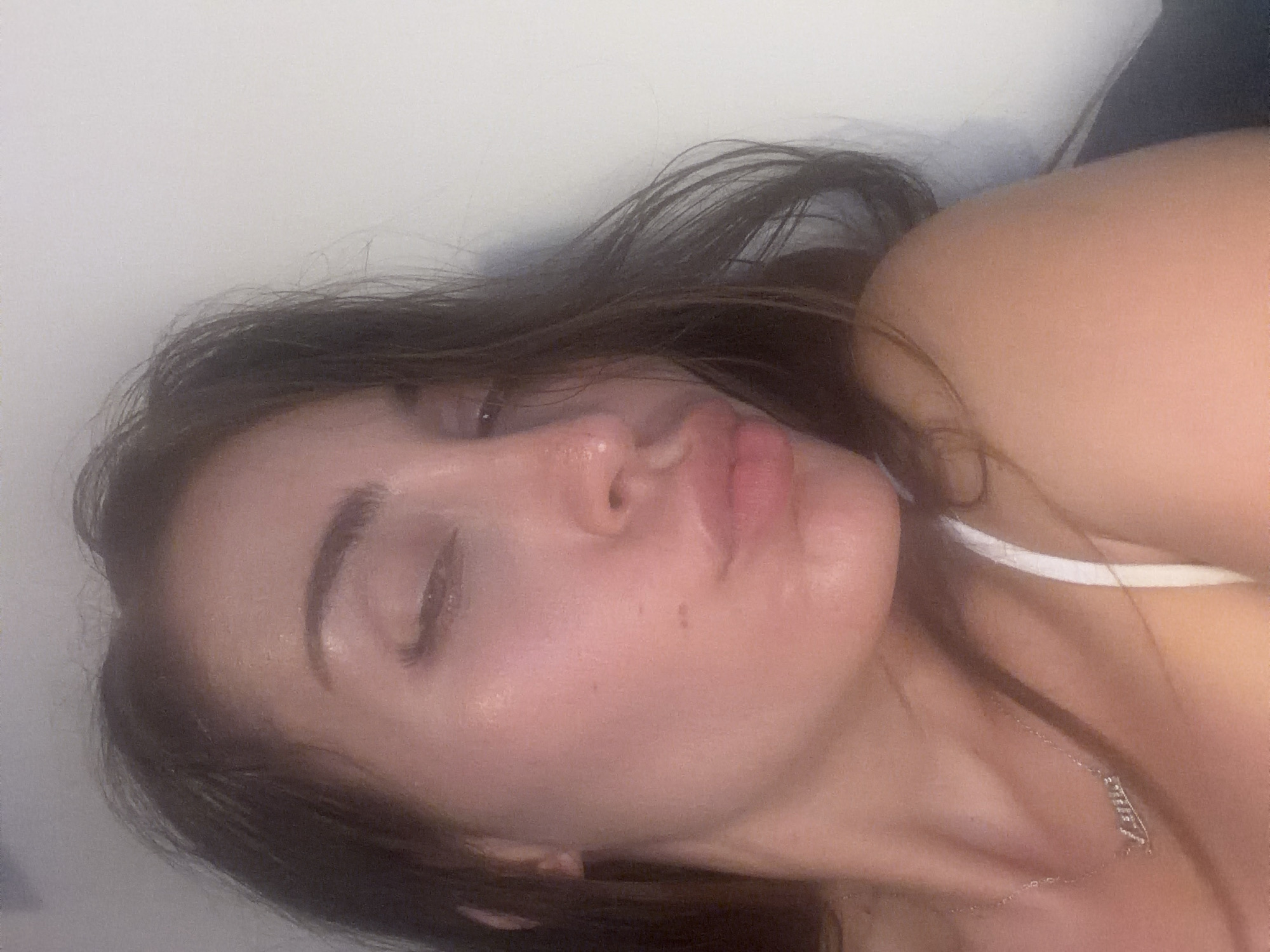 A close-up, chest-up, selfie-style photograph depicts a young Caucasian woman with long, dark brown hair. She is looking downwards and to her left, with her eyes partially closed, and her lips are pursed in a kissing gesture. She is wearing a delicate silver necklace with a pendant that spells out "Lana." A white strap of a top is visible on her right shoulder. The background is plain and light-colored, suggesting an indoor setting with a white wall. The lighting is soft and warm, highlighting the woman's features. The image quality is slightly blurred and grainy. The location is not specified in the image itself, nor can it be determined from visual cues. The image does not provide any information related to the city of Tersefanou, Cyprus.