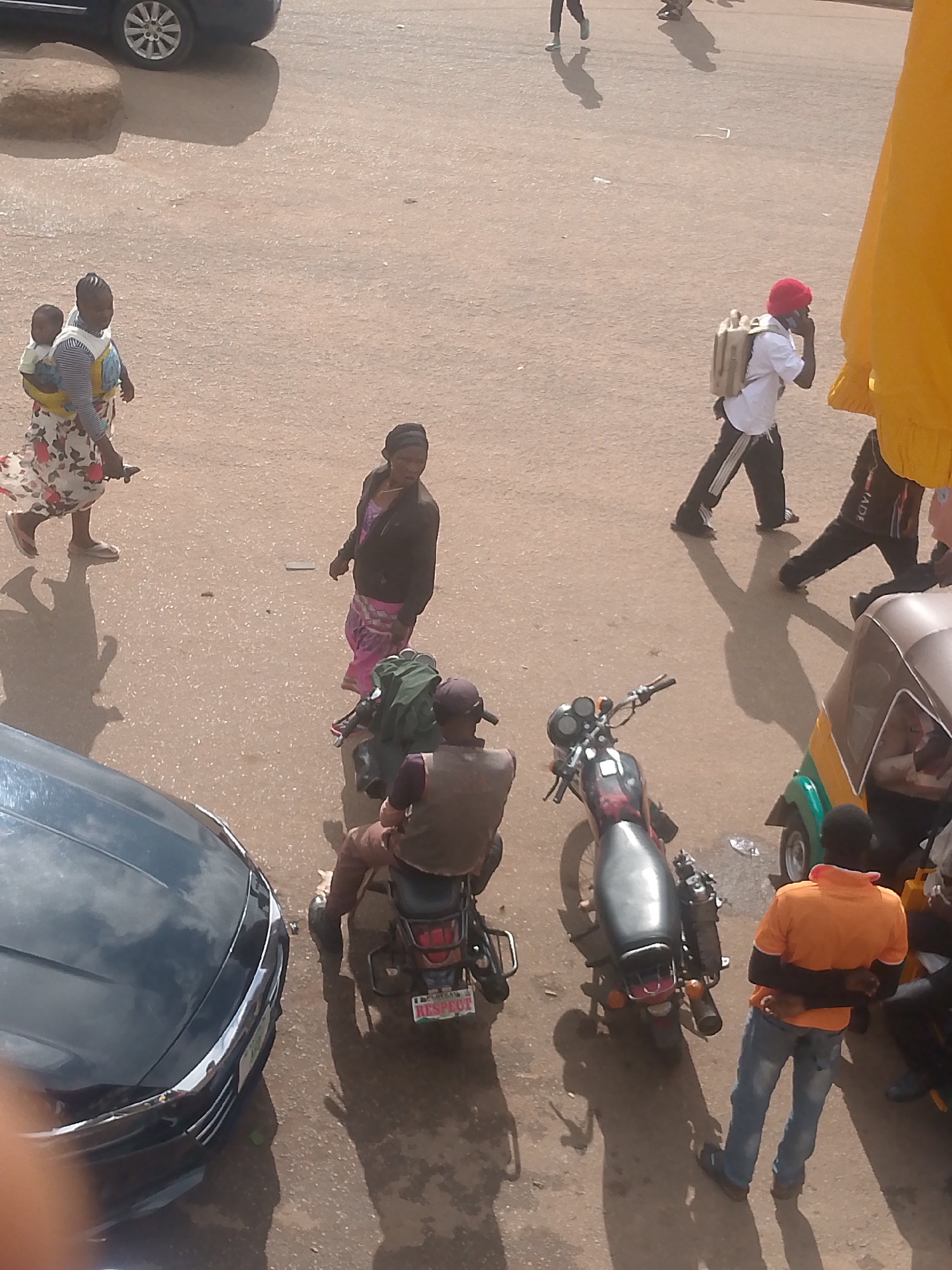 In a scene on a dusty street in Bukuru, Nigeria, several individuals are captured in various activities. A woman with a child on her back walks across the foreground, dressed in a patterned skirt and a striped top. Beside her, another woman is seen walking, wearing a headscarf and a dark jacket over a pink skirt. In the middle ground, a man on a motorcycle is seated, wearing a brown vest and a cap. To his right, a colorful auto-rickshaw is parked. Further back, a man in a white shirt and dark pants with red trim walks while speaking on a cell phone, carrying a backpack. Another man in dark pants and a jacket walks with a similar backpack. The ground is a light brown, dusty surface with scattered debris. In the left foreground, the hood of a dark blue car is visible. The lighting suggests it is daytime, with distinct shadows cast by the people and vehicles.