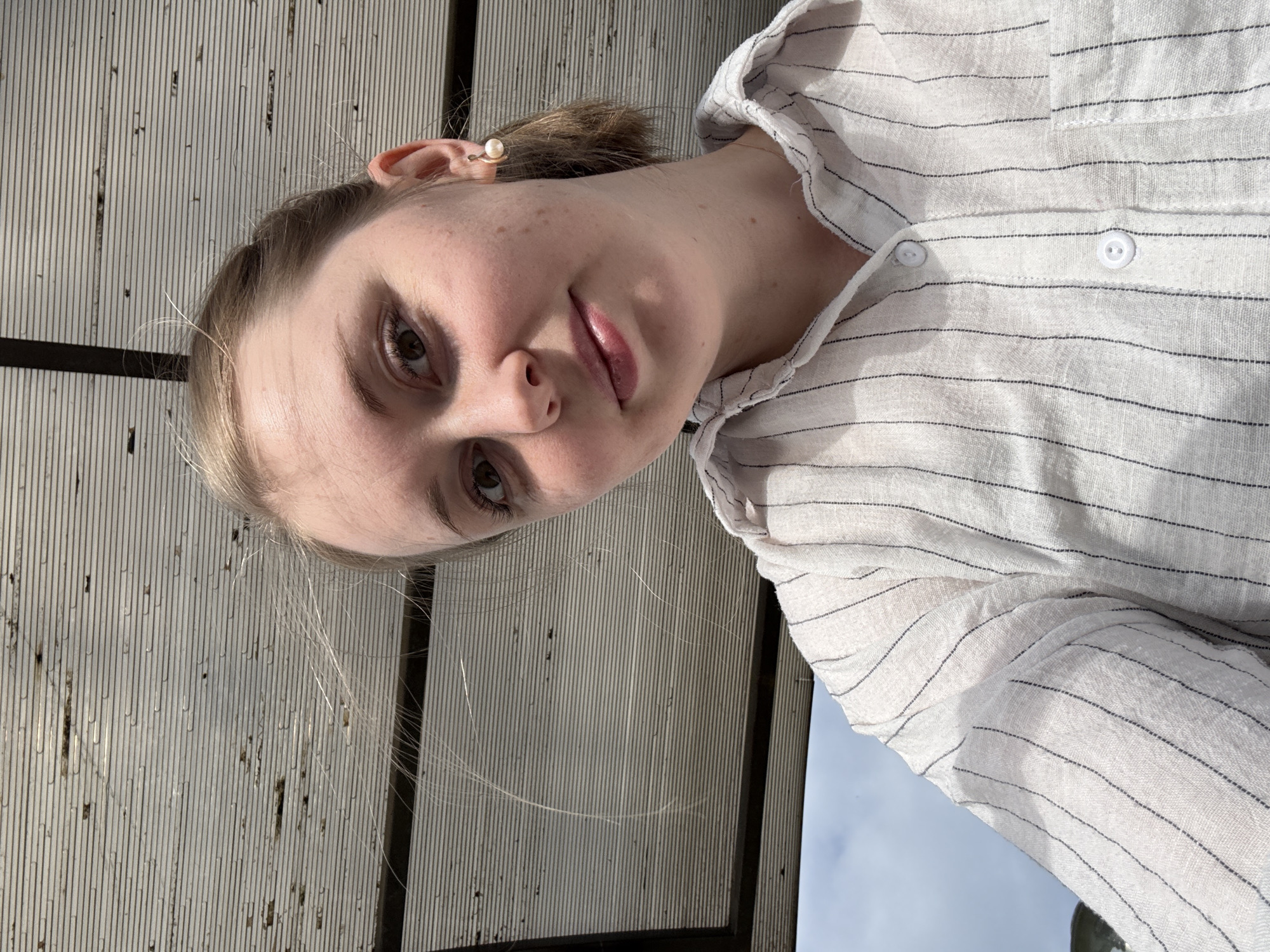 A person with fair skin, brown eyes, and light blonde hair pulled back into a low ponytail is facing the camera at an angle. They are wearing a light-colored, striped button-down shirt. The background consists of a weathered, white, and gray-striped surface. The person is wearing a pearl earring. The location is in the city of Unknown, Unknown.