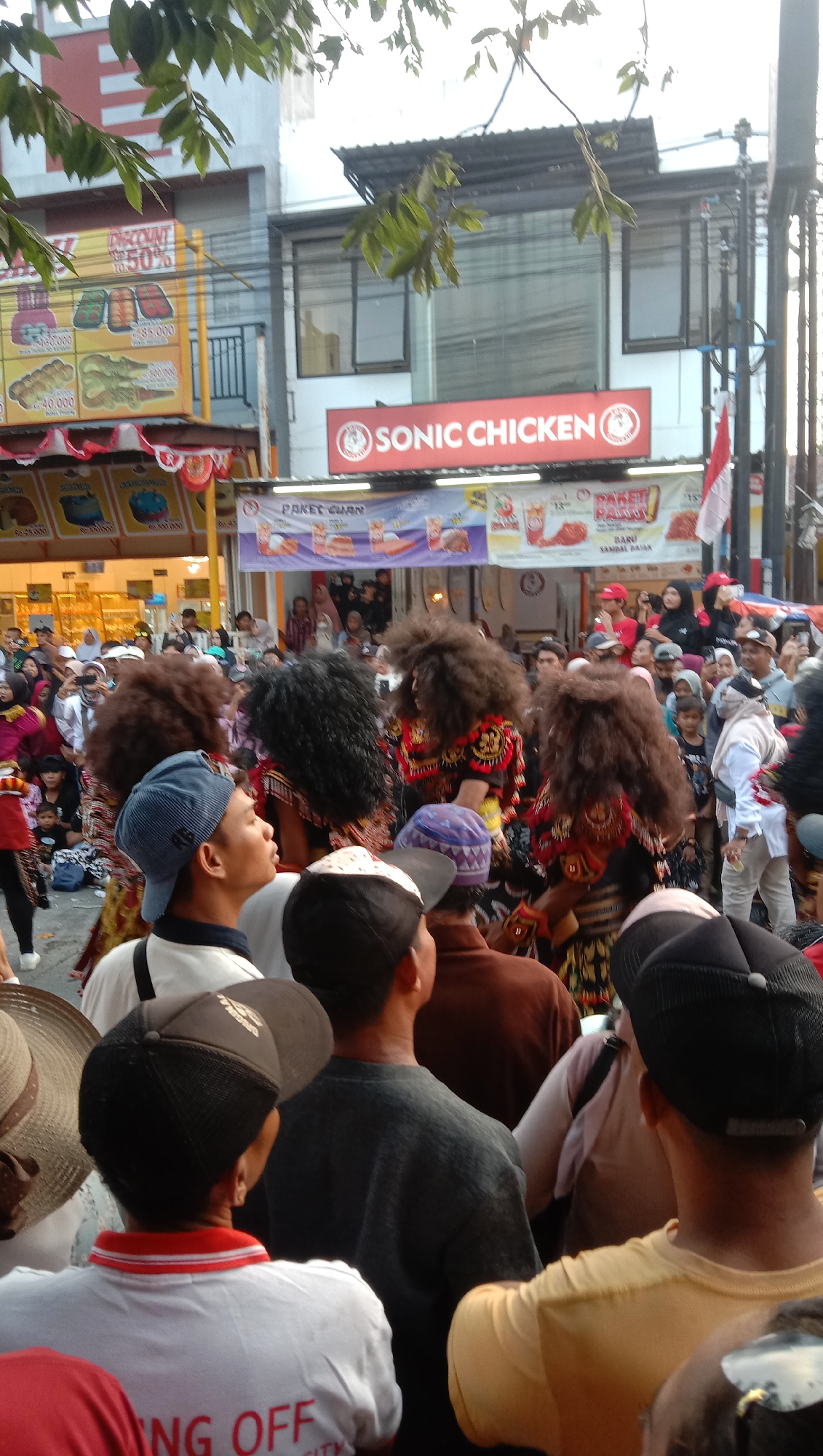 In Jatisawit, Indonesia, an outdoor street scene depicts a gathering of people, likely attending a local event or festival. The foreground is filled with the backs of individuals, many wearing hats and casual clothing. Behind them, a group of performers in elaborate, colorful costumes with large, dark, voluminous wigs are visible, suggesting a traditional dance or performance. Banners displaying "SONIC CHICKEN" and other promotional text are hung above the street, indicating commercial activity. The background shows buildings and awnings, suggesting an urban or semi-urban setting. The overall atmosphere appears festive and crowded.