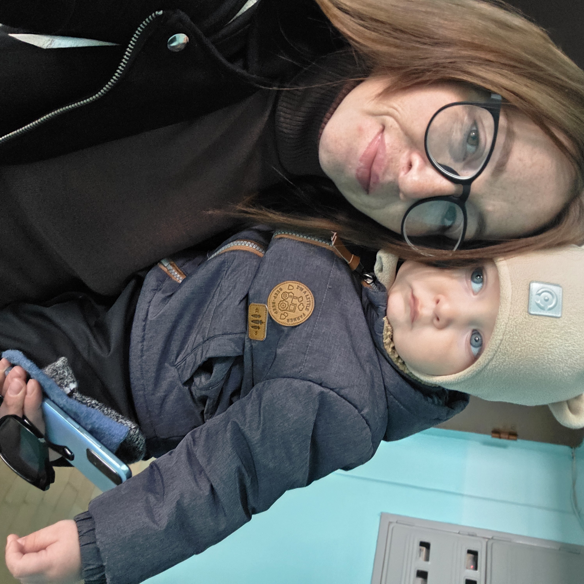 An adult female is depicted holding a young child indoors in Chaikovsky, Russia. The female, positioned on the left, has light brown hair, wears black-framed glasses, a dark turtleneck, and a black jacket, and is looking towards the camera with a slight smile. The child, an infant or toddler with blue eyes, is held in the female's right arm, wearing a light beige beanie with an emblem, a dark blue padded winter jacket with "TRA A LITTLE FARMER BEEP-BEEP" and carrot patches, and dark pants. The child is looking off-camera to the right. A light blue smartphone and dark sunglasses are held in the lower right portion of the frame, likely by the adult. The background features a light blue wall with a visible brown door hinge on the left and a grey electrical panel or utility box with multiple compartments, one exhibiting a red indicator light, on the right.