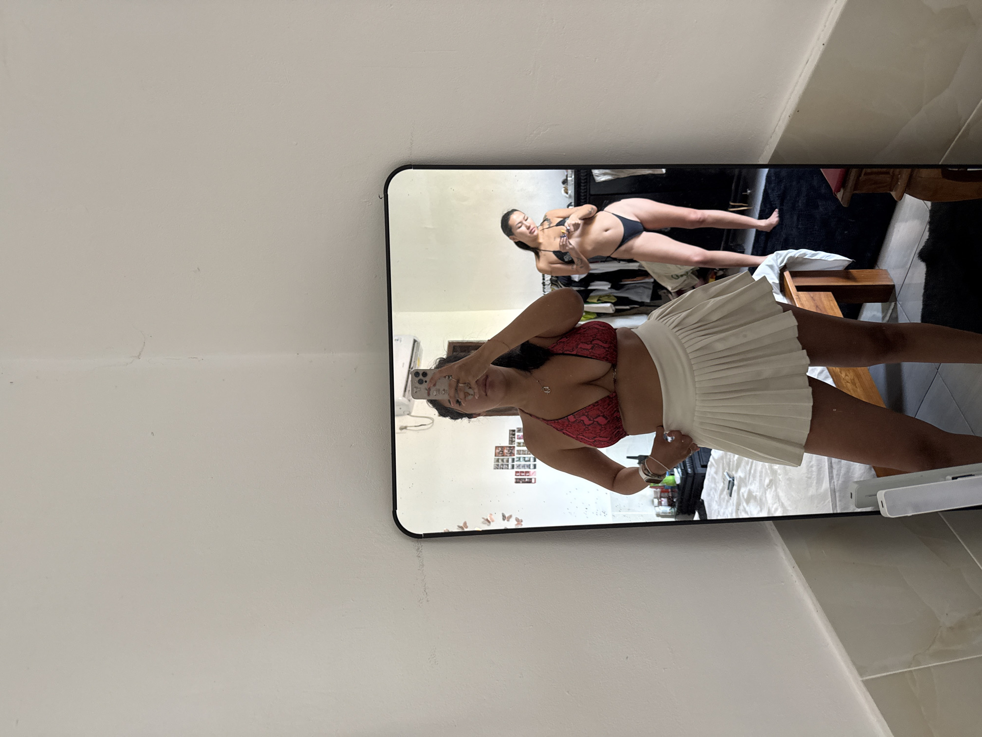 In a bedroom, likely in North Kuta, Indonesia, a woman is taking a mirror selfie. She is wearing a red snake-print bikini top and a white pleated mini-skirt. Her reflection is visible in a large, black-framed mirror, which also captures another woman in the background wearing a black bikini. The room appears to be somewhat cluttered, with clothing racks and a bed visible. The lighting suggests it is daytime, and the overall ambiance is casual and personal.