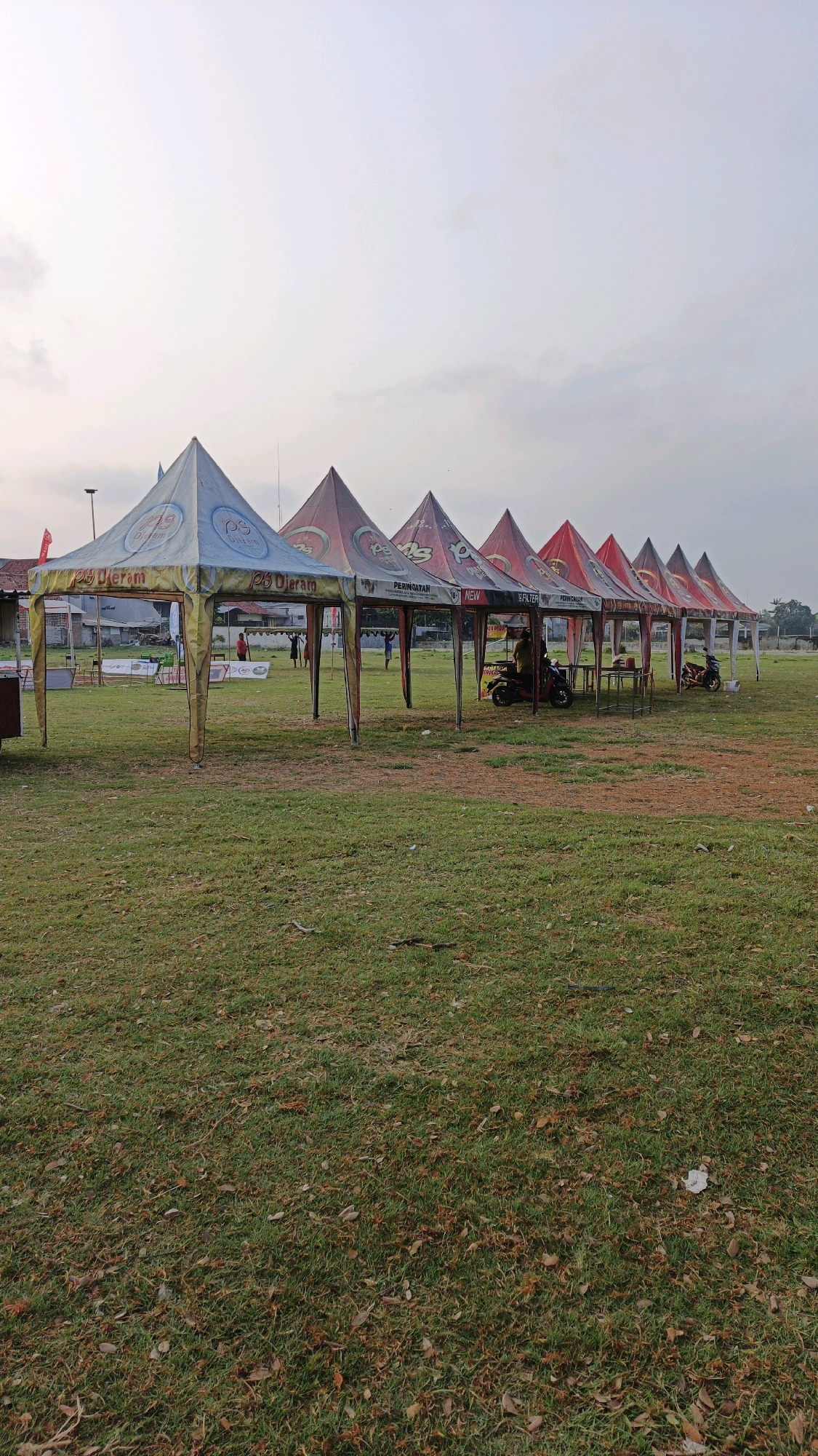 In Boyolangu, Indonesia, a row of six pop-up tents with pointed roofs are set up on a grassy field under a pale sky. The tents are various colors, with the first two predominantly blue and yellow, and the rest red and white, all featuring logos and text. One tent has the text "DJ. jeram" visible. Behind the tents, several individuals are present, some interacting near the stalls and others standing on the grass. A parked motorcycle is visible in front of one of the tents, and another motorcycle is parked further down the row. In the background, there are buildings and trees, suggesting an urban or suburban setting. The ground is covered in green grass with scattered dry leaves.