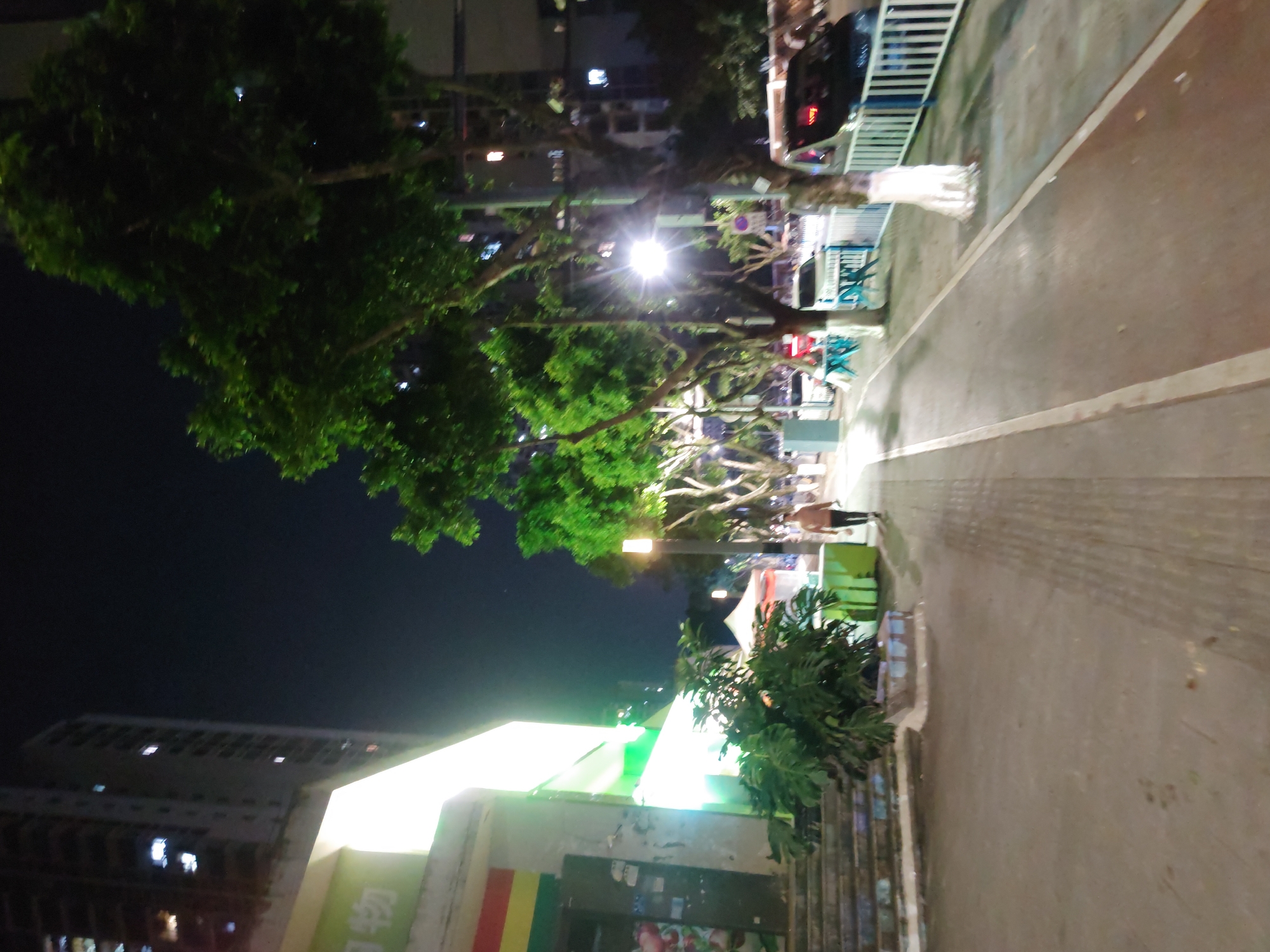 The image depicts an outdoor scene in Chongqing High-tech Zone, China, at night. A pedestrian walkway runs horizontally, with a person visible further down the path. Trees line the walkway, illuminated by streetlights. Buildings are visible in the background, with one structure prominently displaying a bright, lit-up sign. The image is slightly blurred, suggesting movement or low light conditions.