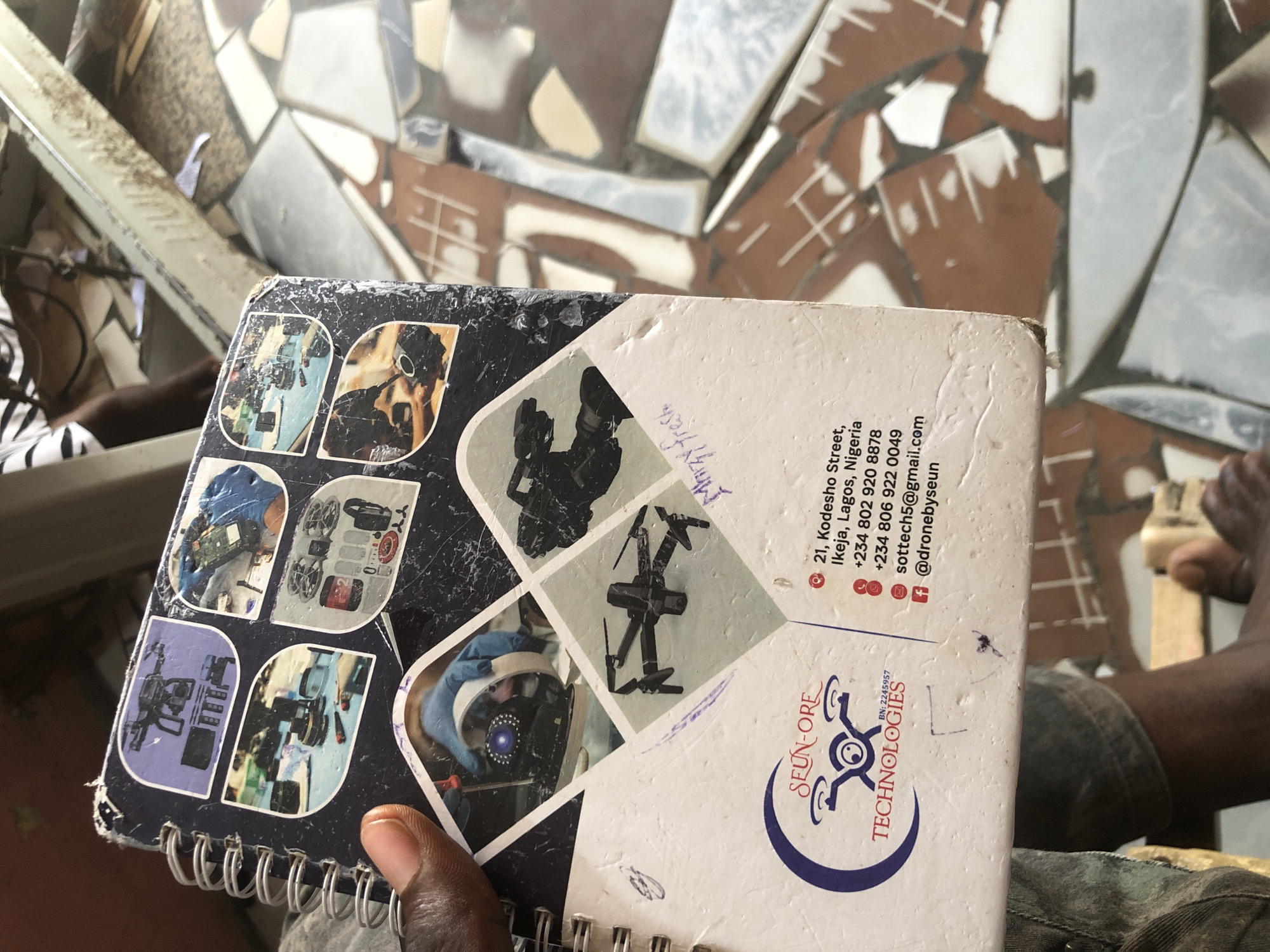 The image shows a close-up of a spiral-bound notebook held by a person's hand. The notebook cover features several small inset images related to drone technology and repairs, including drones, camera equipment, and a close-up of someone working on a drone component. A logo for "SEUN-ORE TECHNOLOGIES" is prominent at the bottom left, along with contact information: an address at "21, Kodesho Street, Ikeja, Lagos, Nigeria," phone numbers, an email address, and a social media handle. A handwritten note in purple ink is visible on the cover. The background suggests an outdoor or semi-outdoor environment with fragmented, tiled surfaces and a blurred, industrial-looking structure. There is no indication of the city of Ado, Nigeria, in the provided image or text.