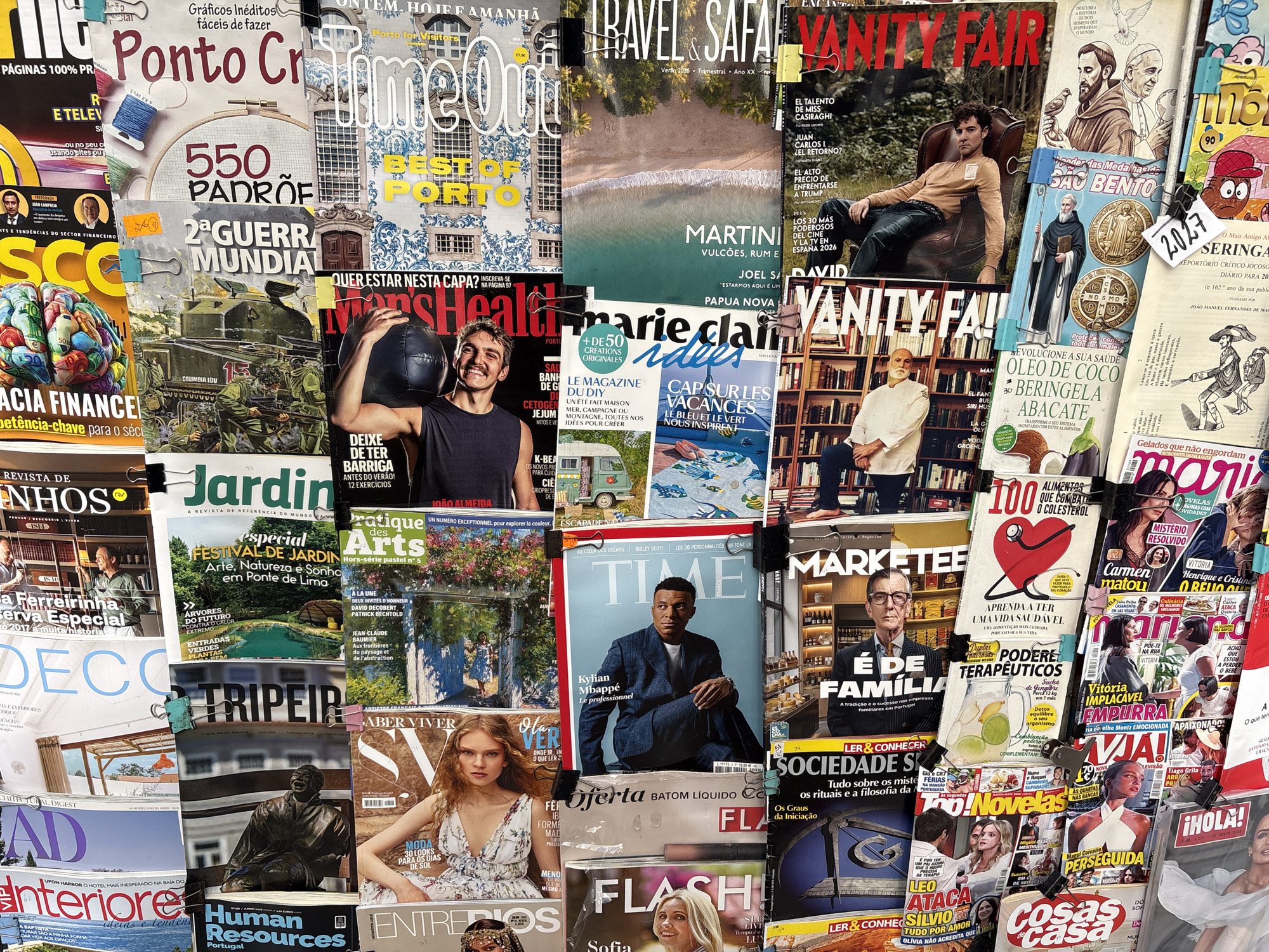 This media file showcases a dense arrangement of magazines, creating a vibrant collage of covers. The magazines cover a wide range of topics, including finance, history, fashion, lifestyle, and celebrity news.
Prominently featured are titles like "Ponto Cr," "2ª GUERRA MUNDIAL," "TIME," "VANITY FAIR," and "¡HOLA!". The magazine covers display diverse subjects. There are portraits of individuals, including a man in a suit on the "TIME" cover, and a woman in a dress on an "¡HOLA!" cover. Other covers feature illustrations, such as those related to religious figures on "SAO BENTO" and historical scenes. There are also covers with visually striking graphics, like the brain depicted on a finance magazine and food-related imagery on health and recipe magazines.
The arrangement suggests a retail or display setting, possibly a newsstand or a market stall in Porto, Portugal, given the presence of Portuguese text and references. The magazines are clipped together, indicating they are part of a collection or display. The overall impression is one of abundance and variety, reflecting the diverse interests of potential readers. The lighting suggests an indoor setting, possibly with natural light contributing to the scene. The overall emotional tone is energetic and informative, driven by the visual appeal and diverse content of the magazines.