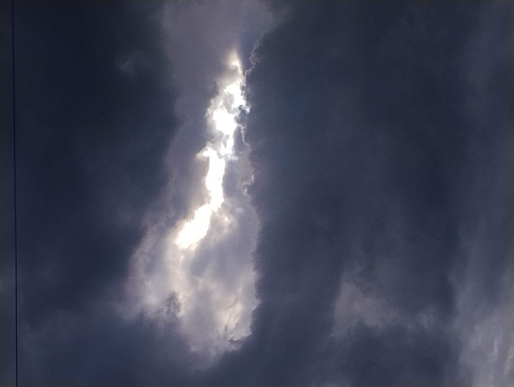 The image captures a dramatic sky filled with dark, heavy clouds, suggesting impending or ongoing stormy weather. A bright, vertical shaft of sunlight pierces through a gap in the clouds, creating a striking contrast between light and shadow. The light illuminates the edges of the clouds, highlighting their texture and form, and casts an ethereal glow. The overall scene evokes a sense of awe, power, and perhaps a touch of mystery. There are no visible people or man-made objects, placing the focus entirely on the natural phenomenon of the sky. The time of day is not explicitly indicated, but the quality of light suggests it could be either dawn or dusk, or during a period of heavy cloud cover when the sun's rays are diffused. The location is unknown.