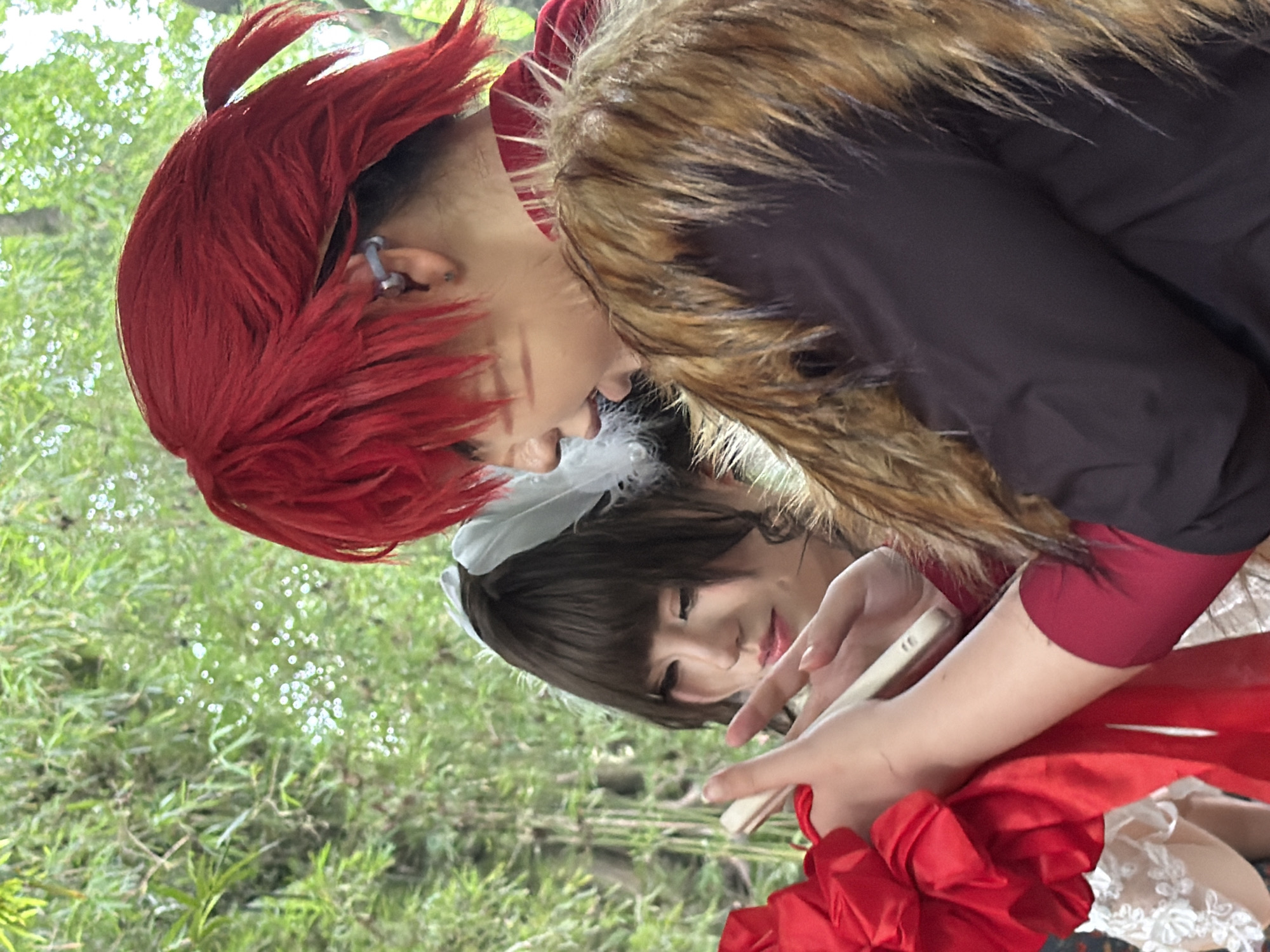 This image depicts two individuals, likely cosplayers, engaged in an outdoor activity amidst lush greenery. The person on the right, with bright red hair and a furry collar, is looking down and appears to be interacting with a handheld device or object. Their face is partially visible, revealing a serious expression. The person on the left has dark hair with a white feather accessory and is also looking downwards, seemingly at the same object. They are wearing a white outfit with lace detailing and a prominent red bow. The background consists of dense foliage, suggesting a natural or park-like setting. The image does not provide any specific information or visual cues that would indicate the location as Yuzhong District, China.