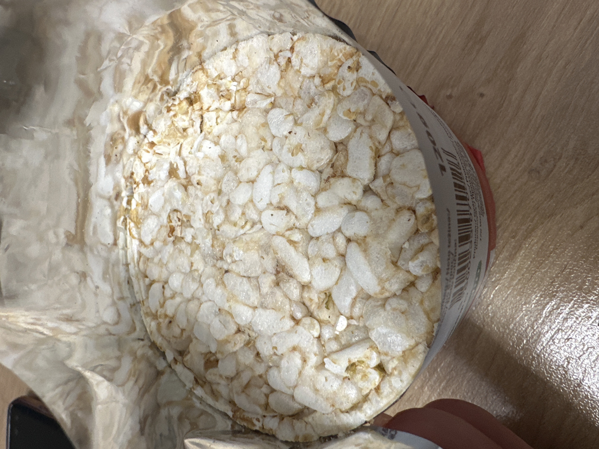An overhead shot captures a package of round rice cakes, likely situated on a wooden surface in Sofia, Bulgaria. The primary subject is a stack of white, puffed rice cakes, visible through a transparent, crinkled plastic wrapper. The rice cakes are composed of individual puffed rice grains, some appearing slightly toasted or browned.
The packaging itself is somewhat translucent, revealing the texture of the product within. A white label with a black barcode and faint, unreadable text is visible on the right side of the packaging, partially obscuring the rice cakes. A human hand, with a visible thumb and finger, holding the package from the bottom right corner, suggesting it is being held or placed on the surface. The fingernails of the visible hand are painted a reddish or pinkish color.
The background consists of a light brown wooden surface, possibly a table or floor, exhibiting a subtle wood grain pattern and some reflective sheen, indicating indoor lighting. The overall scene depicts a simple, everyday moment involving food.