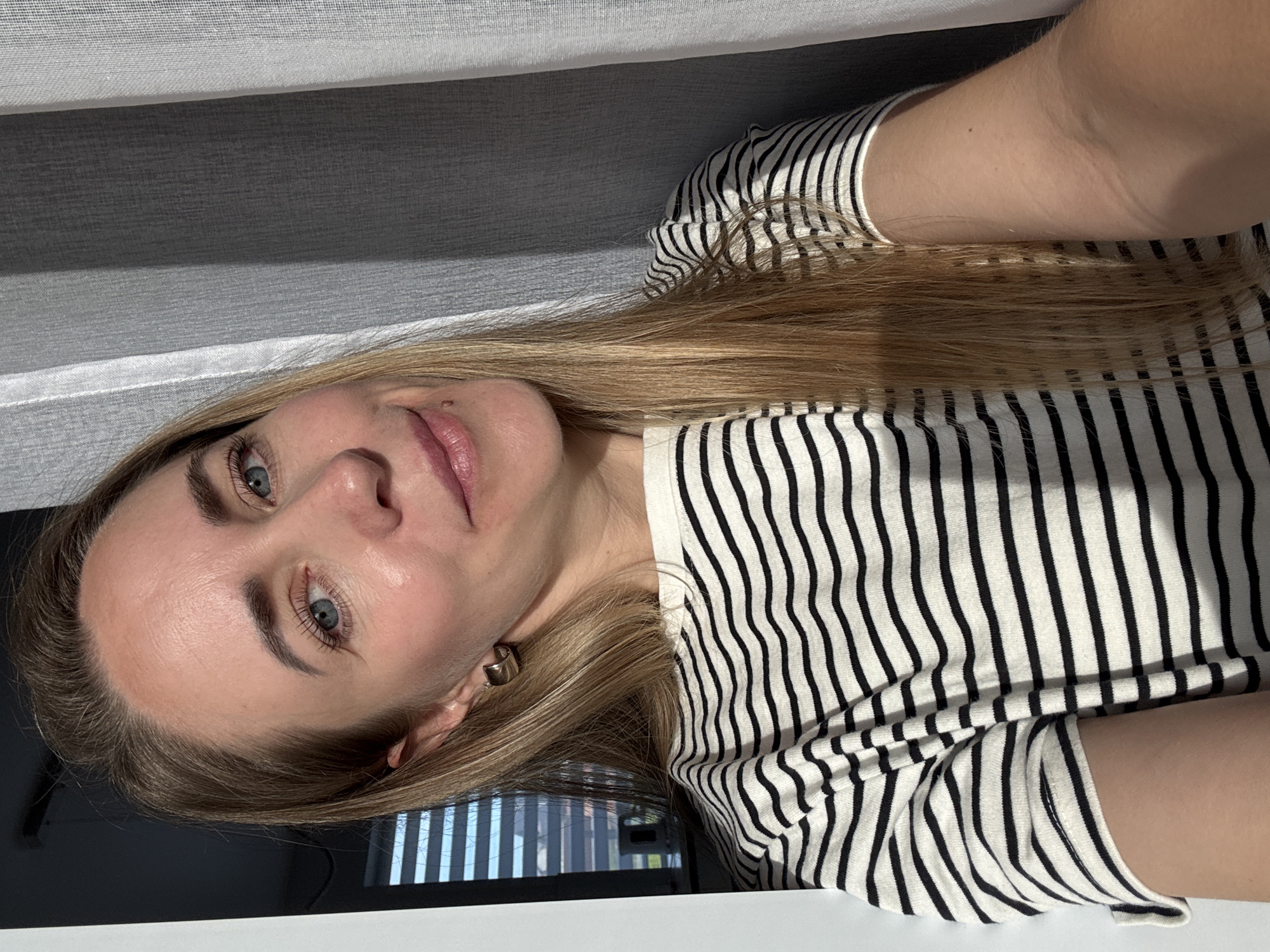 The image captures a head-and-shoulders portrait of a woman, oriented 90 degrees counter-clockwise. The subject is a woman with long, straight blonde hair, blue eyes, and a fair complexion. She has a subtle, pleasant smile and looks directly towards the viewer. A small mole is visible above the right side of her upper lip. She wears a black and white striped long-sleeved top and a small golden hoop earring on her right ear.
The setting is indoors, with the background primarily featuring light and dark grey horizontal blinds or curtains. Below her chin and to the right, a dark, reflective surface, possibly a table or desk, shows reflections of vertical light strips, suggesting a nearby window. The lighting is soft and even, indicative of indirect daylight. The woman's expression conveys a sense of calm and serenity. There is no visible text in the image. This scene is located in Chaikovsky, Russia.