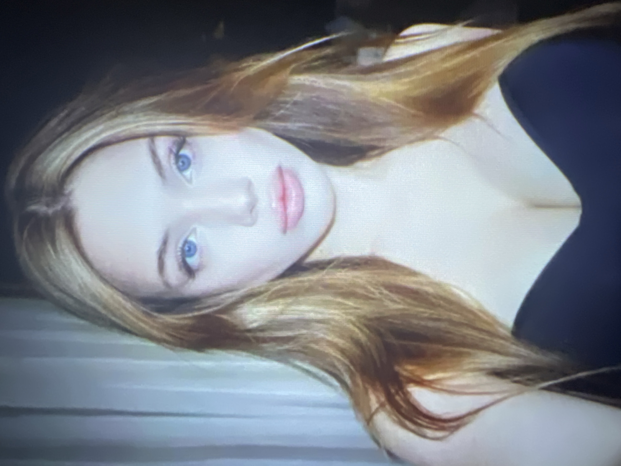 A young woman with long, blonde-brown hair and bright blue eyes is pictured in a close-up portrait. She is wearing a dark, strapless top. The background is blurred, with a suggestion of curtains on the left and darkness on the right. The lighting appears to be soft and diffused. The image quality is slightly grainy, suggesting it may have been taken with a lower-resolution camera or in low light conditions. No specific location in Energetik, Russia is discernible from the image.