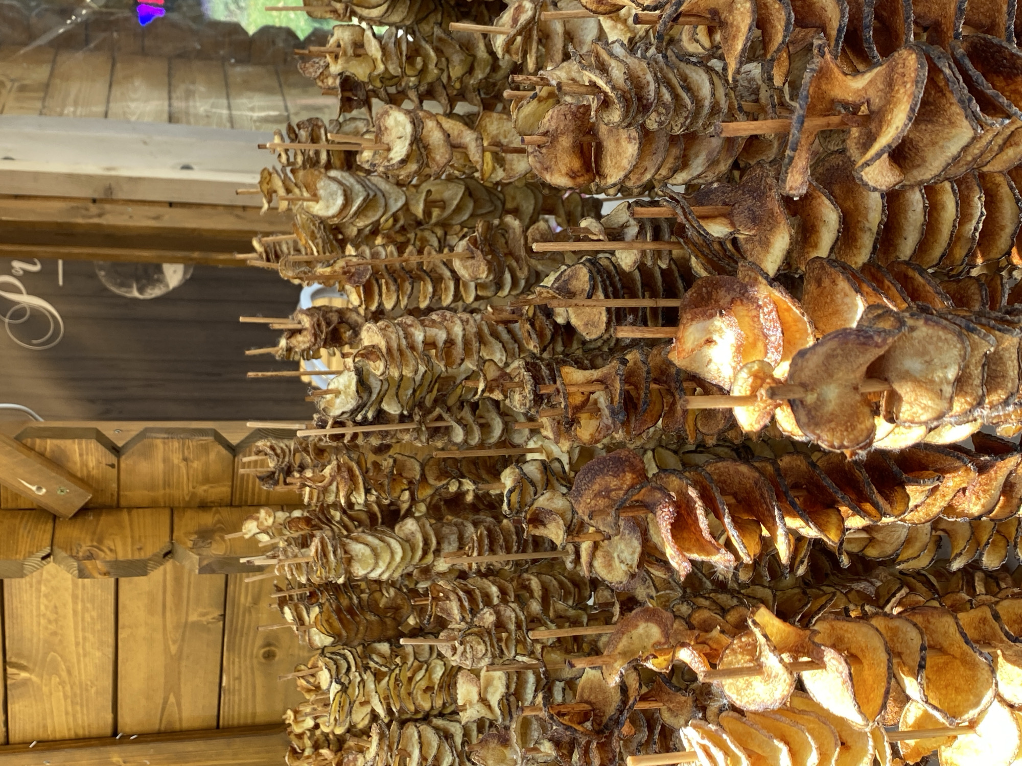 The image shows rows of spiral-cut potato chips skewered on wooden sticks. The chips are displayed under a wooden structure, possibly a market stall or shop. The lighting suggests it is daytime. Based on the spiral potato chips this could possibly be taken at a tourist market in Budapest.