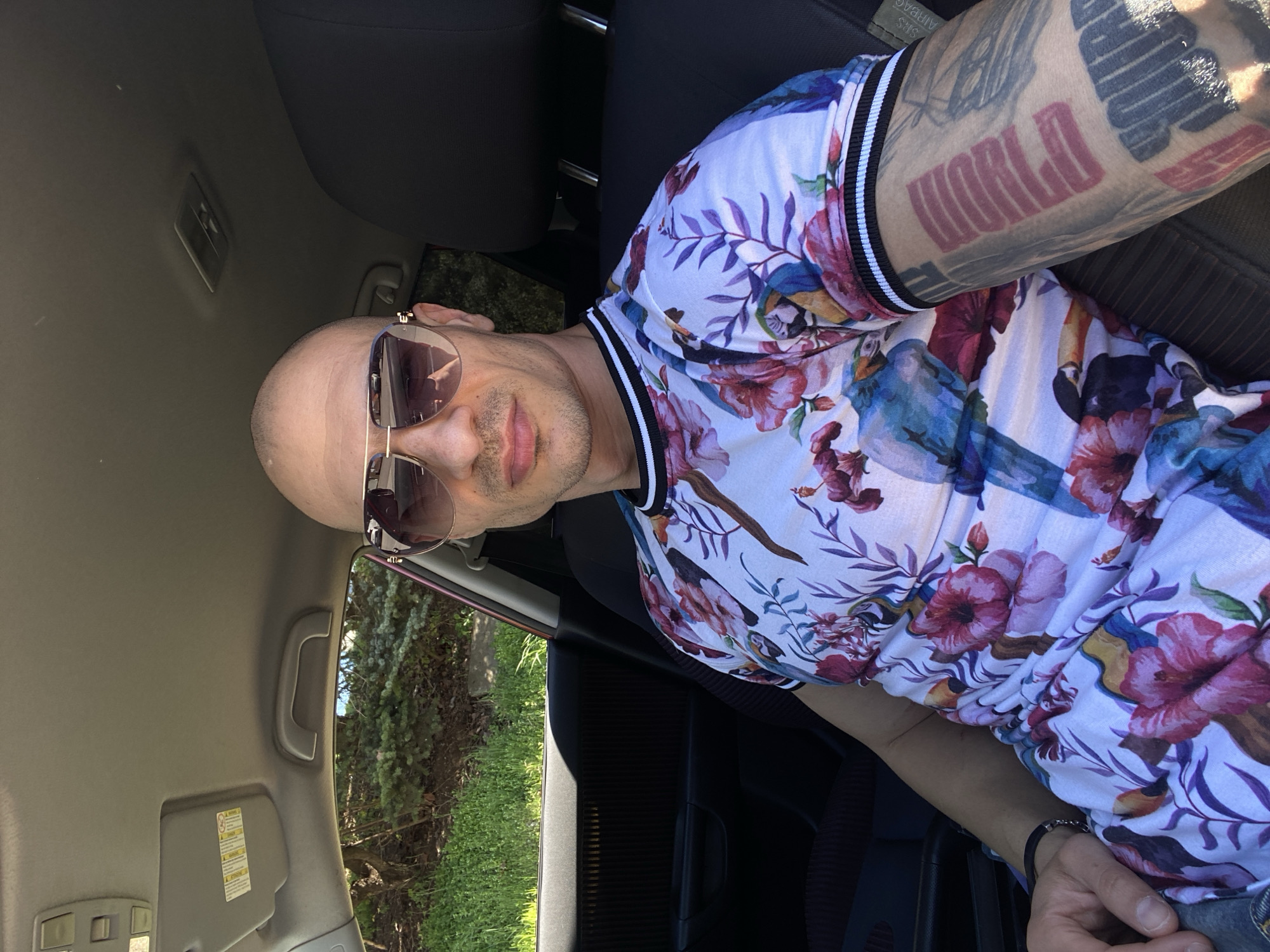 A man is sitting in the driver's seat of a car. He is bald and wearing sunglasses and a white shirt with a floral and bird pattern. He has a tattoo on his right arm that says "WORLD". The interior of the car is visible, including the dashboard, steering wheel, and seats. Outside the car, green foliage is visible. The location is likely not Pleven, Bulgaria.