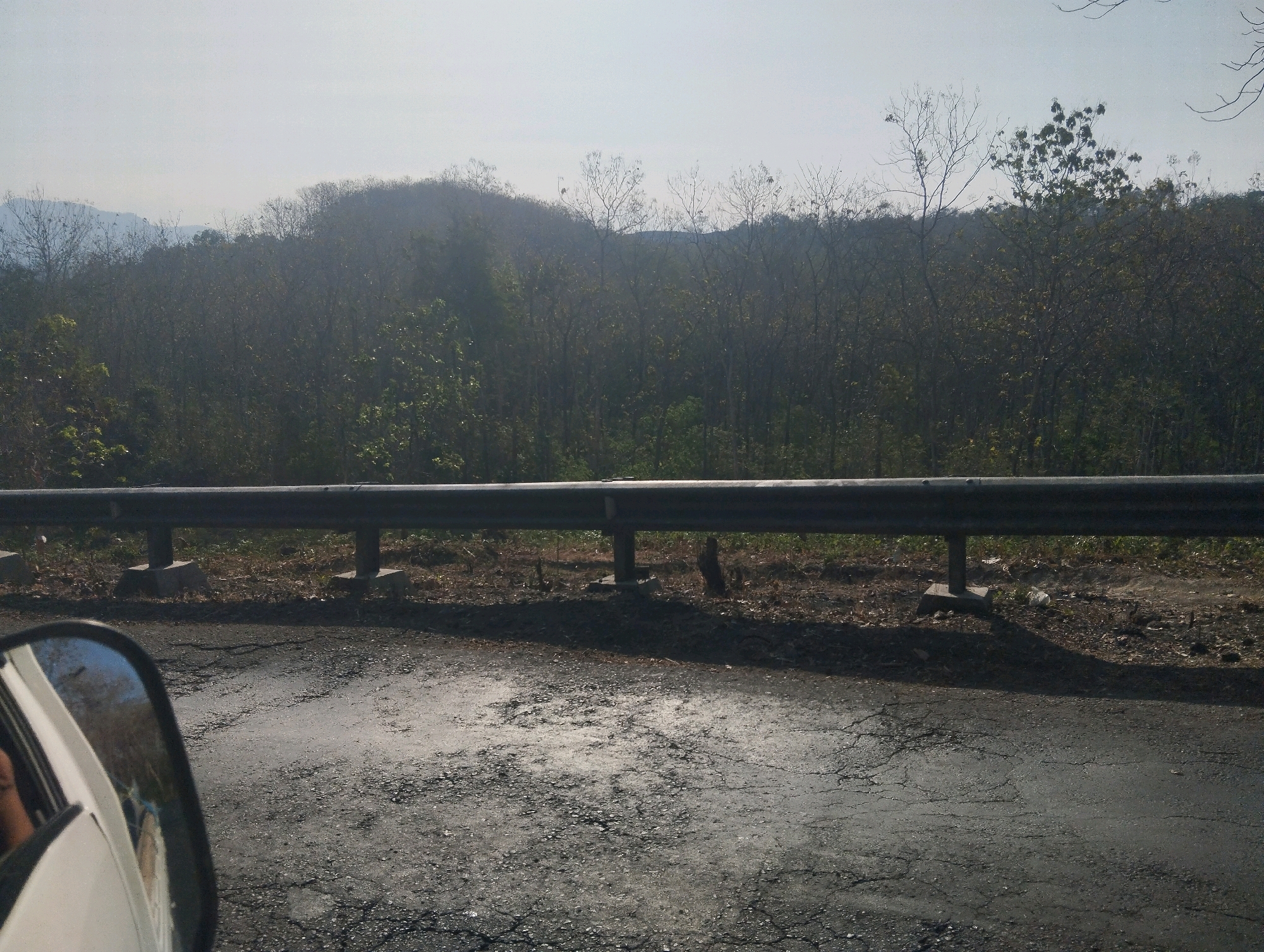 The image captures a roadside scene, likely in Sambi Kerep, Indonesia, from the perspective of a vehicle. In the foreground, a cracked asphalt road surface is visible, with patches of uneven texture. To the left, a portion of a white vehicle's side mirror reflects a glimpse of the road and a person's shoulder within the vehicle. A dark metal guardrail runs horizontally across the middle of the frame, supported by concrete posts. Beyond the guardrail, the ground is covered in dry leaves and sparse vegetation. The background is dominated by a dense forest with a variety of trees, some appearing bare or with minimal foliage, suggesting a dry season. In the distance, hazy hills rise, and the sky is a pale, washed-out blue.