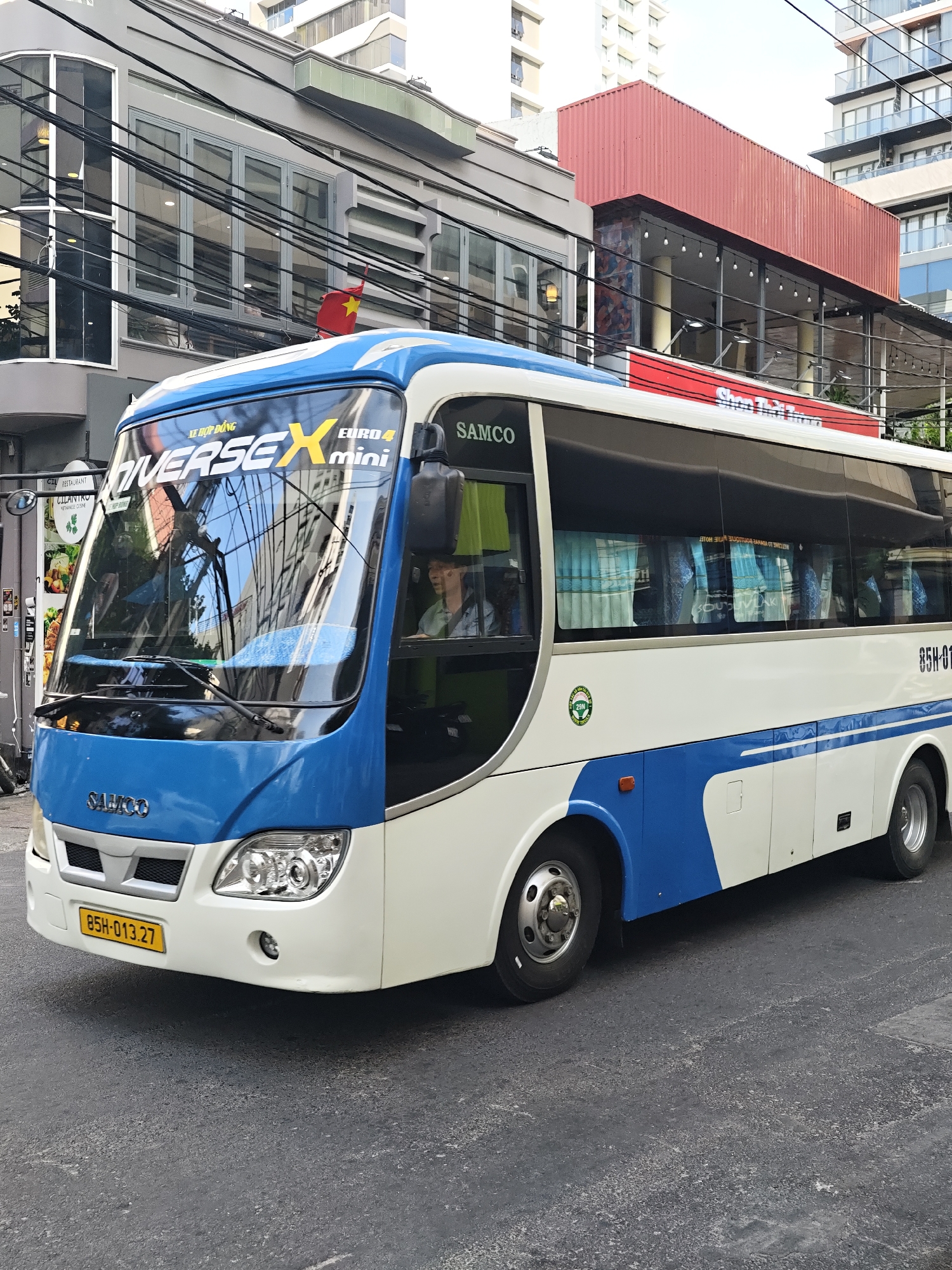 A white and blue bus with the text "DUVERSEX mini EURO 4" on the windshield drives on a paved city street. The bus has a yellow license plate that reads "85H-013.27". The driver is visible through the windshield wearing a light-colored shirt. The bus is passing buildings and overhead electrical wires, characteristic of an urban environment. A Vietnamese flag is visible on one of the buildings. The environment appears to be a sunny day in an urban setting, likely in Vietnam based on the license plate and flag. The bus is the primary subject in the foreground, with the urban background providing context.