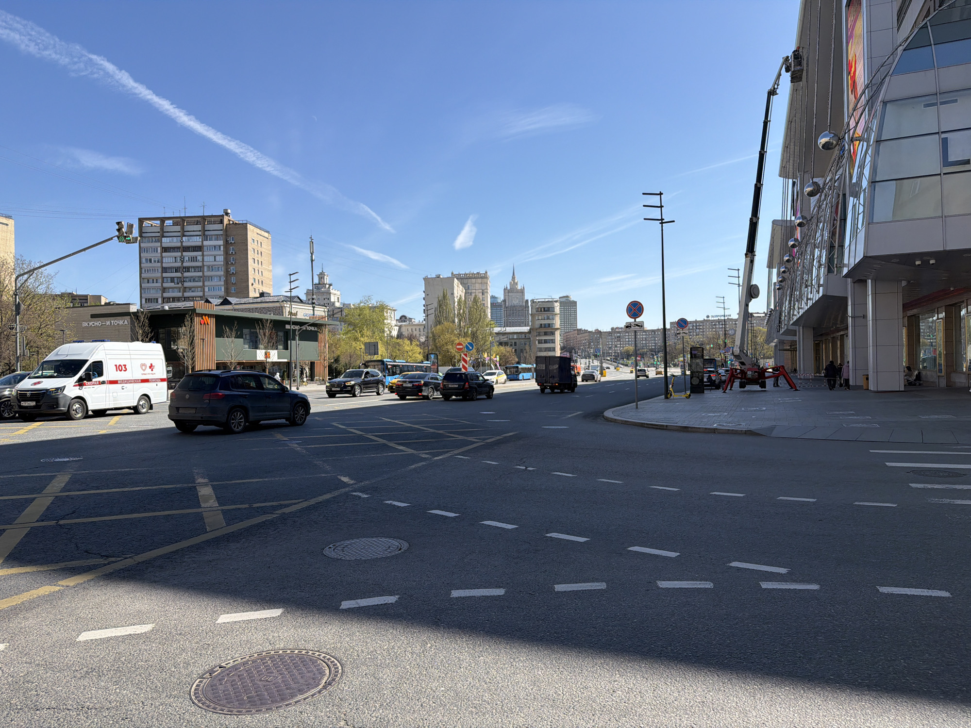 The image captures a vibrant urban intersection in Moscow, Russia, under a clear blue sky on a bright day. The scene is bustling with vehicle traffic and some pedestrian activity.
In the foreground, a multi-lane asphalt road with distinct white and yellow markings, including a 'box junction' cross-hatch, stretches across the frame. A circular manhole cover is visible in the lower left.
To the left, a white ambulance with red markings and the text "103", "МЕДИЦИНСКАЯ ПОМОЩЬ" (Medical Aid), and "СКОРАЯ" (Ambulance) is seen at a standstill, possibly at a traffic light. Behind it, a building with "ВКУСНО — И ТОЧКА" (Vkusno i Tochka – "Delicious. Full Stop."), the rebranded former McDonald's, is visible. Several other cars, including dark sedans and SUVs, are present on the road, with some moving through the intersection.
The background features a mix of architectural styles, from older residential buildings to modern structures. A tall, distinct Soviet-era skyscraper with a spire is visible in the distant center. A traffic light with green signals indicates active traffic flow. A sign for "ГАЗПРОМБАНК" (Gazprombank) can be seen further down the street.
On the right side of the image, a large, modern building with a glass facade and reflective decorative spheres hanging from its exterior dominates. A worker in a basket attached to an extended boom lift is engaged in activity, possibly maintenance or installation, on the upper levels of this building. A few people are visible on the sidewalk below.
The time of day appears to be late morning or early afternoon, indicated by the clear sunshine and visible shadows. The weather is fair with minimal cloud cover, save for a few faint contrails. The overall impression is that of a typical, active city street environment.
