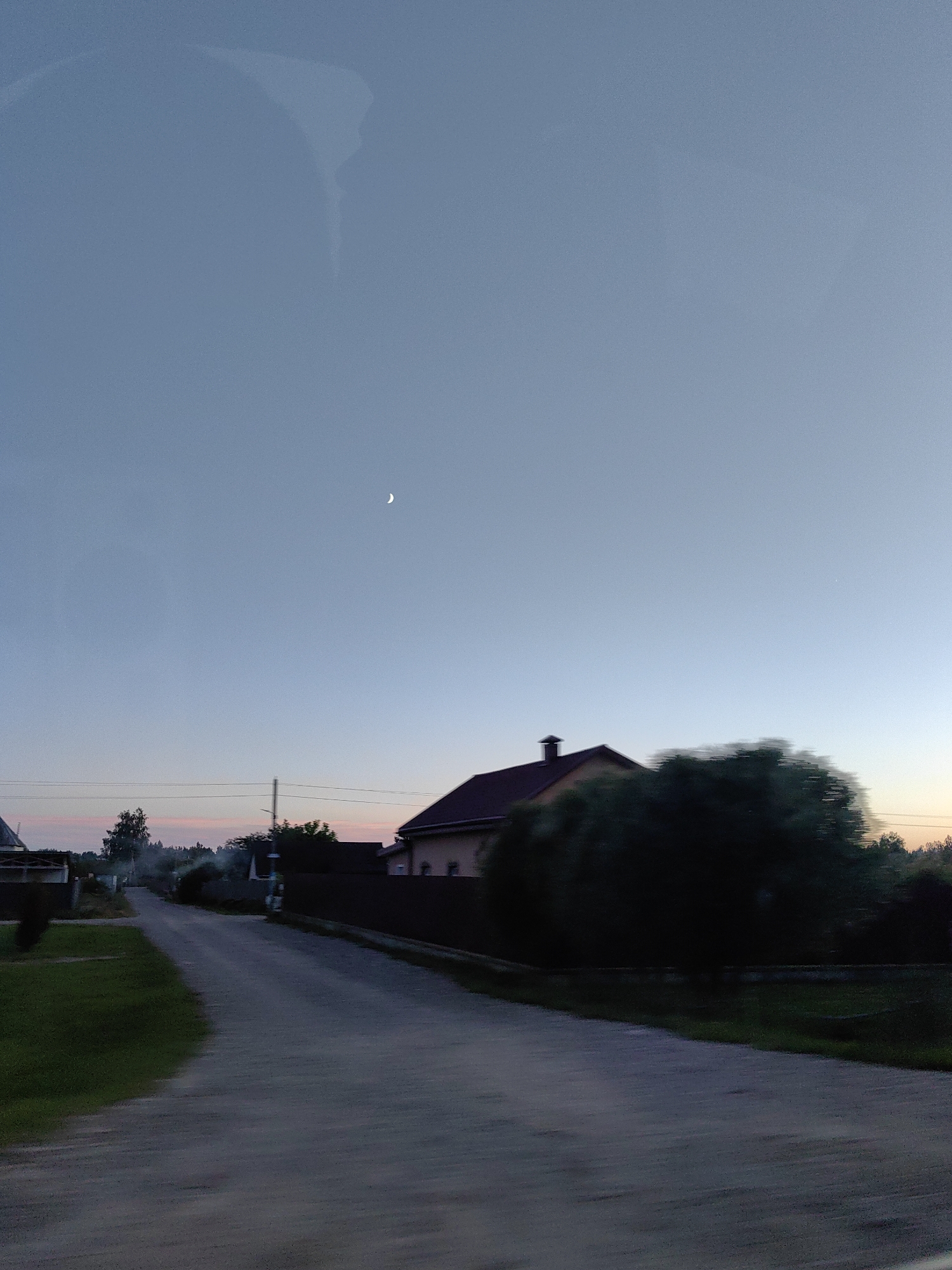 The image captures a serene twilight scene in a rural Belarusian village. A crescent moon is visible in the pale blue sky, which gradually transitions to hues of pink and orange near the horizon, suggesting the aftermath of sunset. The foreground features a paved road, leading the eye towards a cluster of houses and trees. The motion blur indicates that the photo was taken from a moving vehicle. One house, with a dark red roof and a chimney, is prominently featured on the right side of the road, partially obscured by a large, dark green tree. Other houses and fences can be seen further down the road and on the left side, blending into the dusky landscape. Smoke is rising from behind the trees in the distance, hinting at domestic activity. The overall atmosphere is peaceful and quiet, characteristic of a rural settlement in Belarus during the evening.
