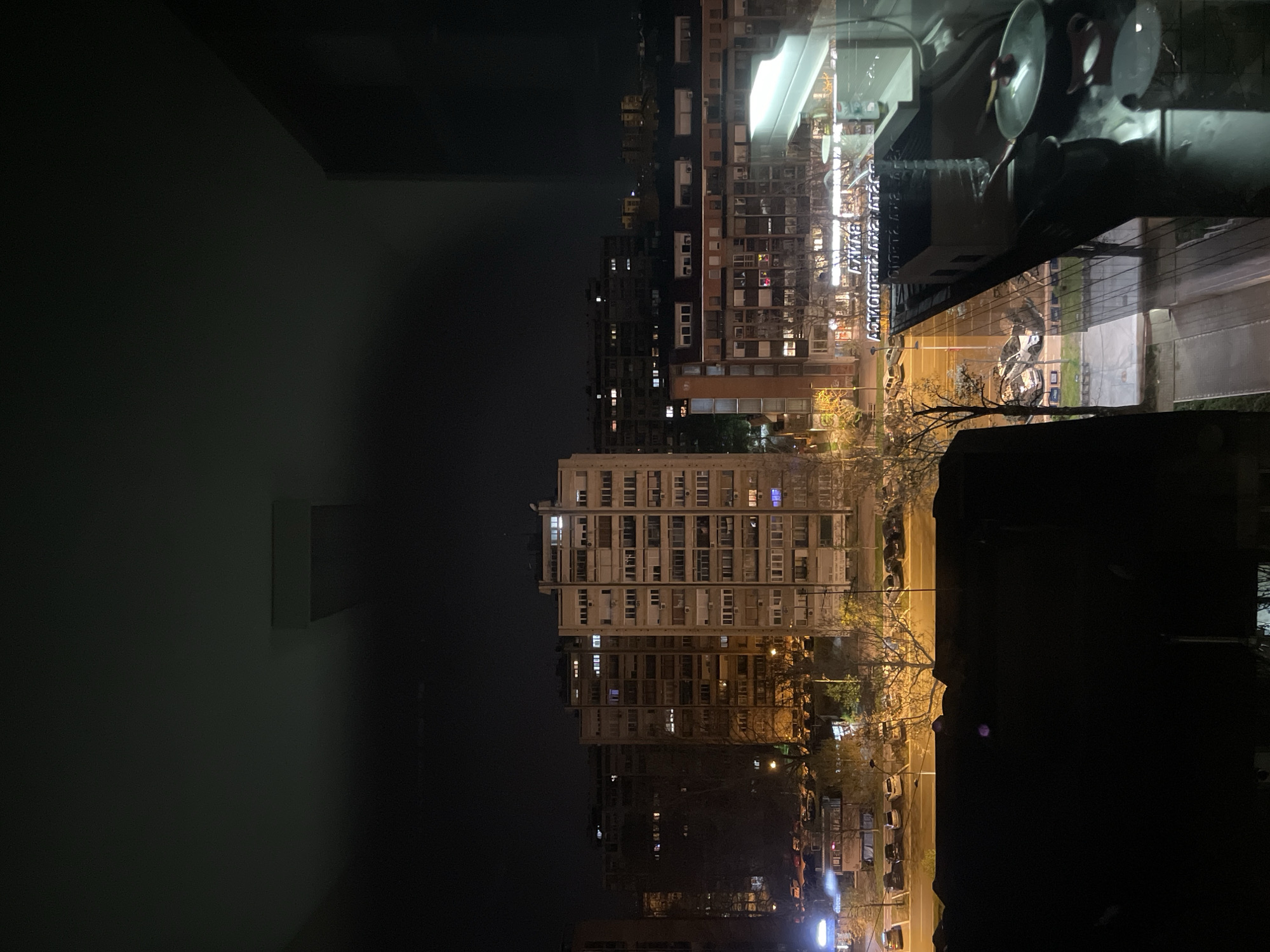 The image shows a nighttime urban scene in Novi Sad, Serbia, viewed through a window. The window reflects indoor objects, including a dish rack with dishes. Outside, multiple apartment buildings of varying heights are visible, illuminated by streetlights and lights from the buildings. A street runs along the base of the buildings with parked cars along the side. Bare trees line the street. The sky is dark. The scene suggests a residential neighborhood at night.