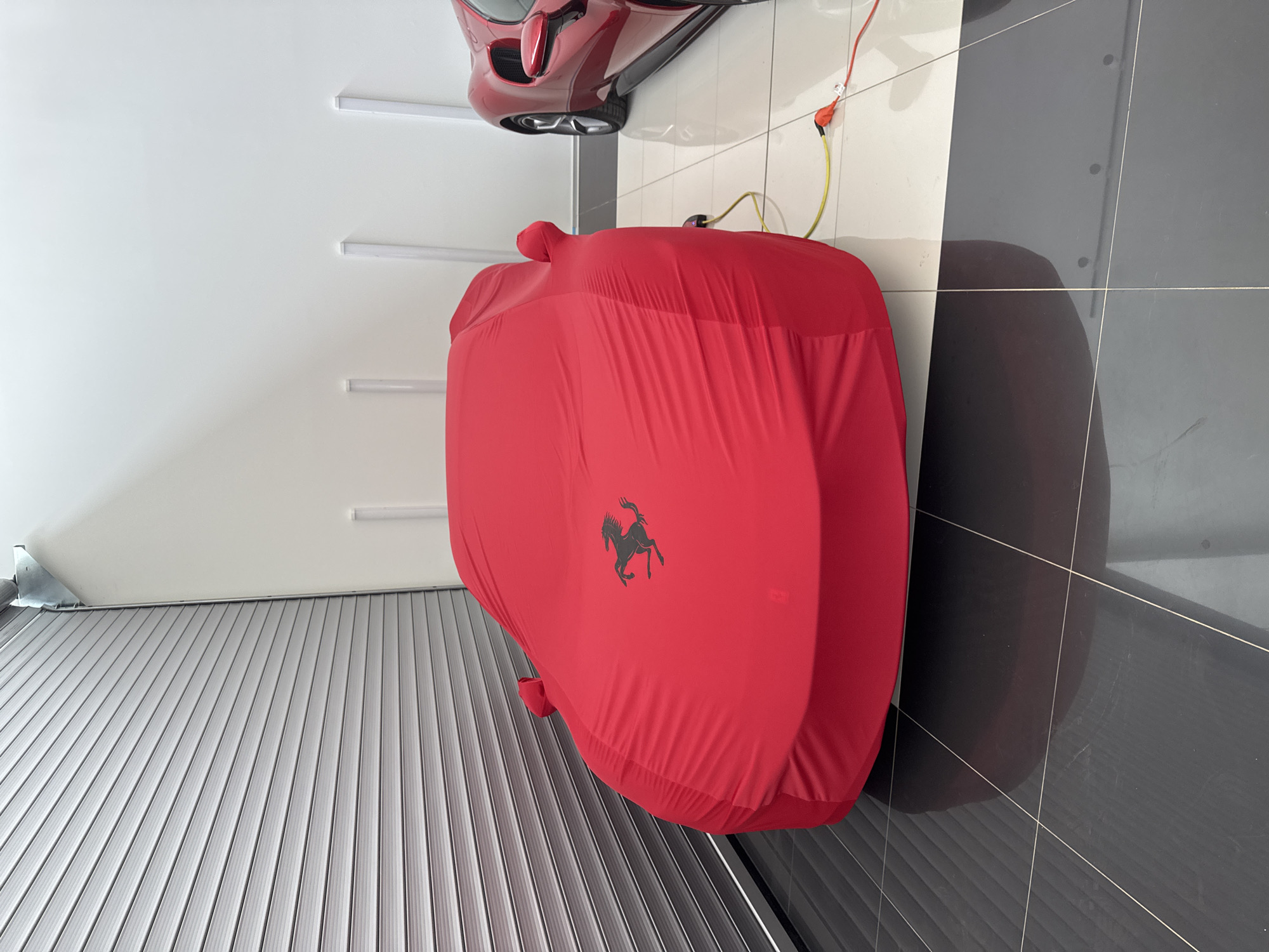 The vertically oriented image displays a meticulously kept indoor space, characteristic of a private garage or luxury car showroom in Sofia, Bulgaria.
The primary subject is a car completely enveloped in a vibrant red custom car cover, resting on a tiled floor. A distinct black silhouette of the Ferrari prancing horse logo is emblazoned on the side of this red cover, indicating the brand of the vehicle beneath.
Above and to the right of the covered car, a second red sports car, likely also a Ferrari, is partially visible. This vehicle appears to be mounted high on a wall or ceiling, showcasing its rear underside and a wheel. A red and yellow electrical cable, possibly connected to a battery charger, runs from near this elevated car towards a power outlet on the white wall.
The flooring features large, glossy tiles laid in a pattern of alternating black and white sections. The walls are predominantly white, with several long, rectangular fluorescent light fixtures illuminating the space. To the bottom left of the image, a gray, horizontally ribbed metal rolling shutter or garage door is partially visible. The scene is devoid of people and conveys a sense of careful preservation and display of high-performance automobiles.