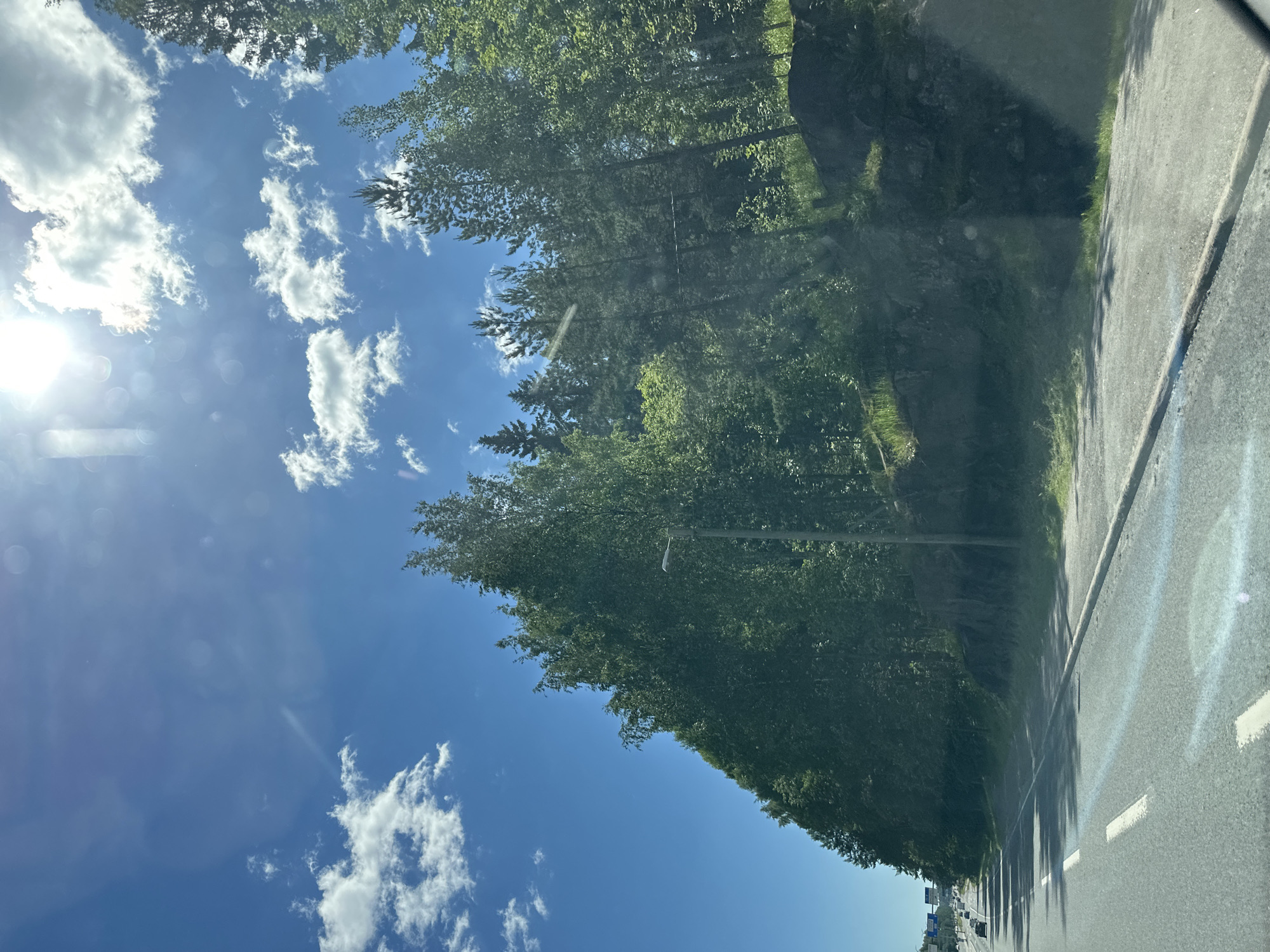An outdoor, eye-level, long shot captures a clear blue sky with sparse, fluffy white clouds. The sun is brightly shining in the upper right quadrant of the image. A dense forest of green trees lines the right side of a paved road, which stretches into the distance on the left. The road has visible white lane markings. A few cars are driving on the road in the distance. There is a lamppost on the right side of the road, partially obscured by trees. A rocky embankment is visible behind the trees.