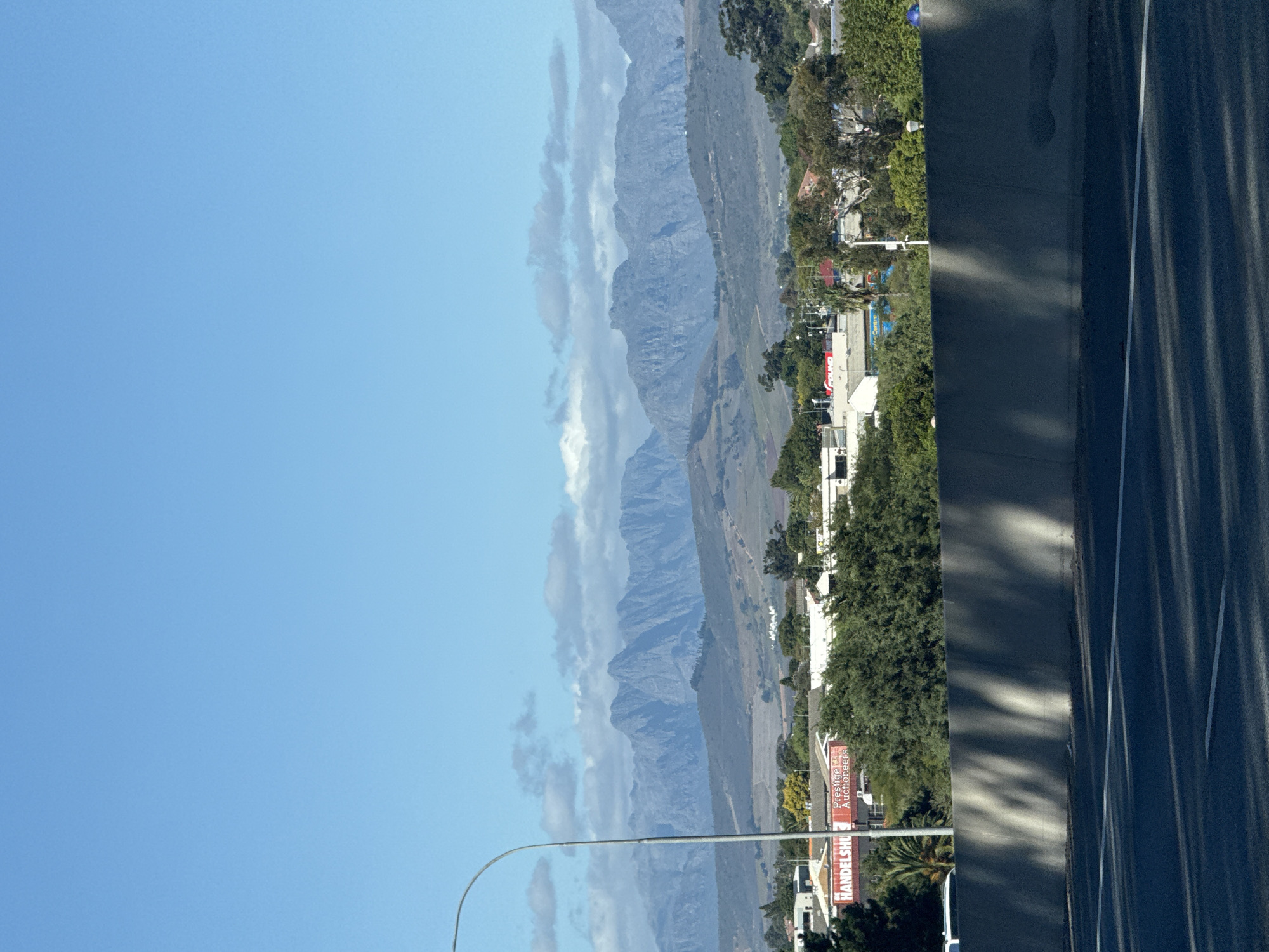 This media file captures a panoramic outdoor scene, likely taken from a vehicle or roadside viewpoint, looking towards a prominent mountain range. The image, oriented vertically, displays a clear blue sky with some scattered white clouds, particularly around the mountain peaks, indicating good weather conditions during the day.
In the mid-distance, a long, rugged chain of greyish-blue mountains stretches across the horizon, partially veiled by light clouds. The mountains feature distinct ridges and valleys. Below the mountains, the landscape transitions into a mix of green fields, trees, and scattered buildings.
In the foreground and mid-ground on the right side of the frame, there is a dark asphalt road or highway with clear white lane markings. Long, diagonal shadows are cast across the road surface and an adjacent barrier or wall, suggesting bright sunlight and possibly morning or late afternoon.
On the left side of the road, a commercial or residential area is visible, featuring green trees and buildings. Several red signs with white text are discernable. One sign clearly reads "Prestige Auctioneers" with "HANDELSHU" (likely part of "Handelshuis," meaning trading house) partially visible below it. Another red sign further back appears to have the text "TIENERS" or similar. A curved street light arm extends from the bottom of the frame towards the center.
The scene depicts a blend of urban infrastructure and natural landscape, consistent with areas near Bellville, South Africa, which offers views of surrounding mountains. No specific human activity or identifiable individuals are visible. The atmosphere is calm and clear.
