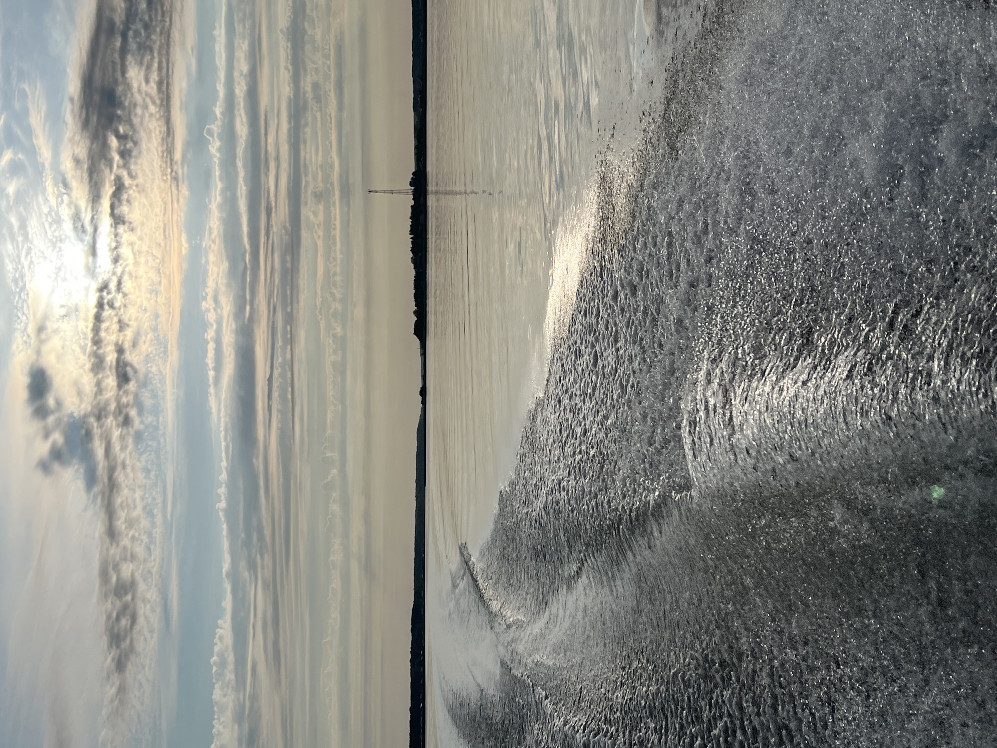 A body of water, likely a river or lake, is depicted with a boat's wake prominently in the foreground, extending from the left side of the image and creating frothy, white patterns on the water's surface. The wake is illuminated by sunlight, highlighting its texture and movement. The water is calm beyond the wake, reflecting the sky and distant landscape. In the distance, a silhouetted shoreline is visible, dotted with trees and some structures. A tall, slender tower, possibly a radio or observation tower, stands out against the horizon. The sky is filled with a layer of clouds that appear to be thin and streaky, with patches of sunlight breaking through, suggesting it is either sunrise or sunset. The overall impression is one of a serene, natural environment, possibly filmed from a moving vessel. The location is not explicitly shown, but it is claimed to be in Novopichugovo, Russia. There are no people visible in the image.