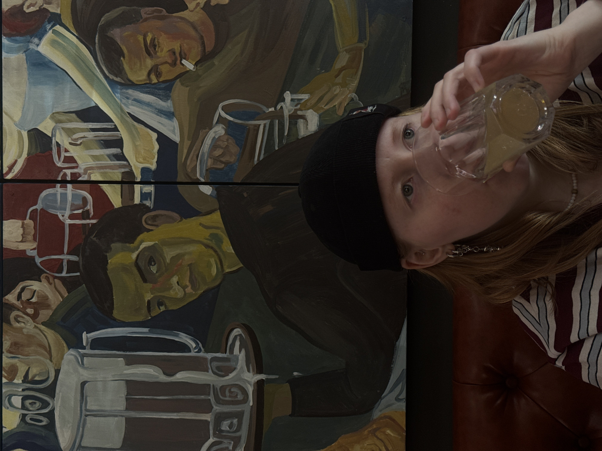 A person wearing a black beanie and a striped shirt is drinking from a glass, in front of a painting depicting men with beer mugs. The painting is reminiscent of socialist realism. The person is sitting on a reddish-brown leather-like surface. Based on other information, the location may be in the city of Perm, Russia.