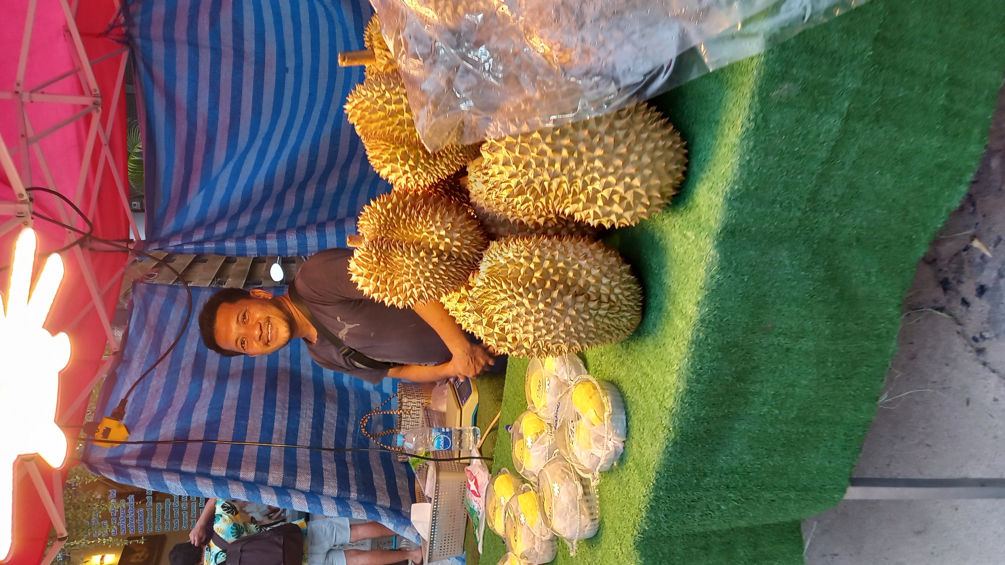 This image depicts an outdoor market stall in ตำบลเกาะพะงัน, Thailand. The primary subject is a male vendor, appearing to be in his 30s or 40s, with short dark hair, a beard, and a mustache, positioned behind a table. He wears a dark grey t-shirt with a light emblem and a black shoulder bag, and is smiling towards the camera.
The table, covered with green artificial turf, displays various durian products. On the right, a pile of approximately five whole, spiky, golden-yellow durians is visible. To the left, seven clear plastic containers hold peeled, pale yellow durian flesh segments. Behind these containers, a clear plastic water bottle labeled "Sada" is present, alongside a woven basket and a white digital weighing scale. A large, clear plastic bag partially filled with an obscured product is on the far right of the table.
The stall is illuminated by two bright, bare light bulbs suspended from a red and pink canopy structure. The rear of the stall is covered by blue and white horizontally striped tarpaulins. In the upper left background, a brown wooden sign, partially obscured, displays text in Thai and English, listing services such as "Traditional Thai M", "Oil Massage", "Foot Massage", "Back and Shou", "Coconut Oil M", "Thai Aroma M", and "Mas".
On the far left, a person with short hair, wearing a multi-colored floral shirt and light shorts with a black backpack, is partially visible from the back, interacting with an unseen item near the wooden sign. The ground beneath the stall is a grey concrete surface.