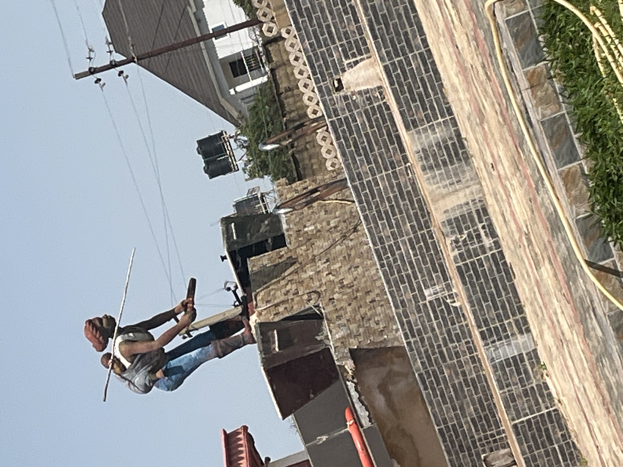 The image depicts a statue of a woman carrying a baby on her back, with a long stick or pole balanced on her shoulders. She is wearing blue trousers, brown boots, and a white top, with a red head covering. The statue stands atop a structure made of brick and stone, with a red roof visible to the left. In the background, a white building with a dark roof and a power pole with electrical wires are visible. The scene appears to be outdoors under a clear sky. The statue's placement and the surrounding architecture suggest it is a monument or landmark, possibly in an urban setting. Without further information, the precise location within Jos, Nigeria, cannot be definitively determined from the image alone. However, the architectural style of the buildings and the presence of a power pole are consistent with urban areas in Nigeria.