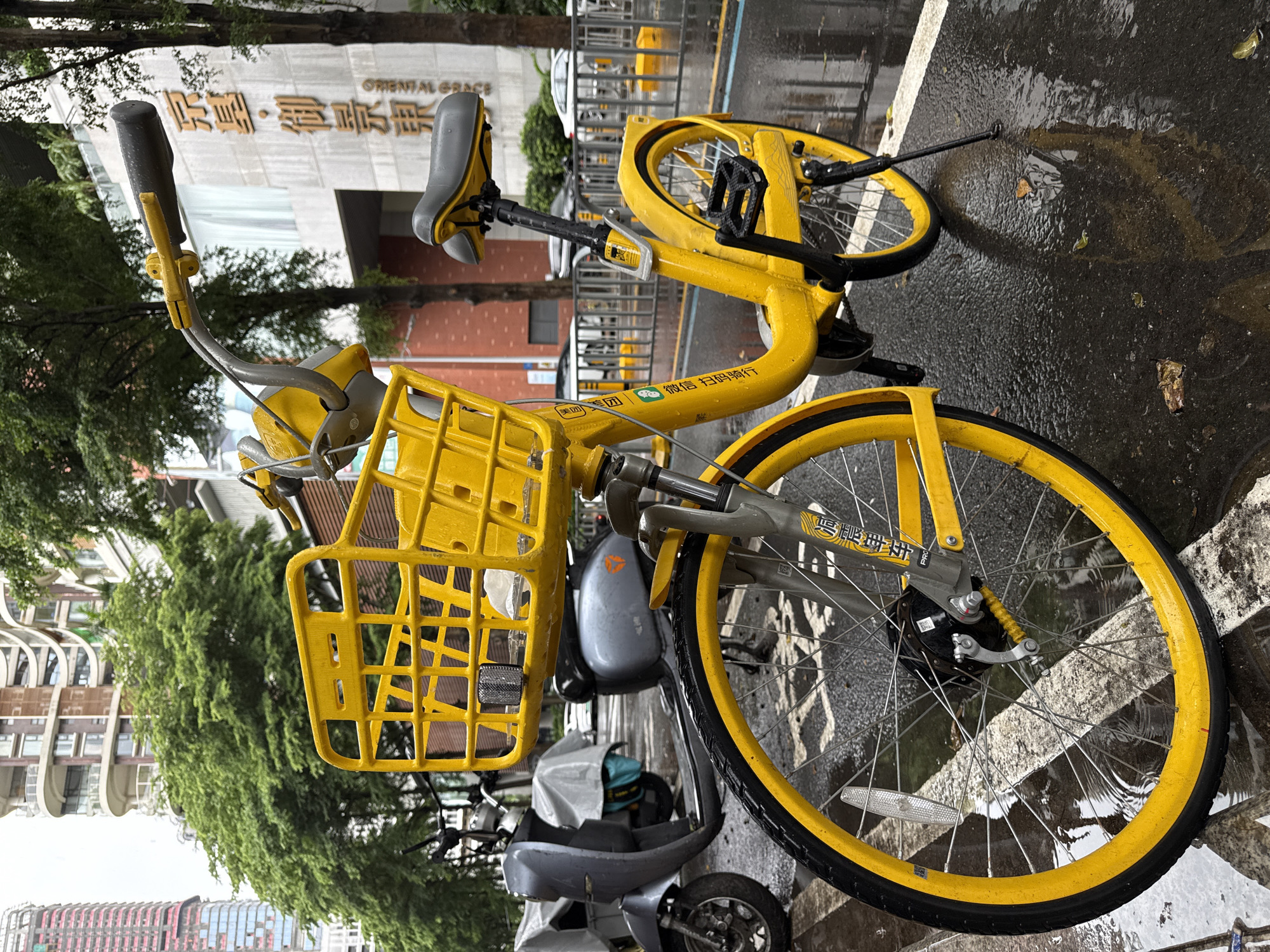 A bright yellow bicycle with a yellow basket is parked on a wet street in Nanshan District, China. The street is wet from rain, reflecting the bicycle and surrounding buildings. In the background, there are modern buildings with glass facades and trees lining the street. A gray scooter is parked behind the bicycle. The bicycle has a black seat and black pedals. There are Chinese characters on the frame of the bicycle. The overall scene depicts an urban street environment after a rainfall.