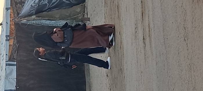Two individuals are depicted walking on an unpaved, light-colored dirt ground in Khan Yunis, Palestinian Territory. They are moving from right to left within the frame. The person in the foreground, an adult female, wears a brown abaya and a black hijab, carrying a dark shoulder bag on her right side, and black and white athletic shoes. The person behind her, a younger female, wears a black jacket, dark trousers, and white athletic shoes. Her hair is in a ponytail, and she carries a light-colored shoulder bag on her left side. In the background, temporary structures consistent with tents or shelters are visible, featuring dark fabric walls and light-colored tarp roofs with dark striping and patterns.