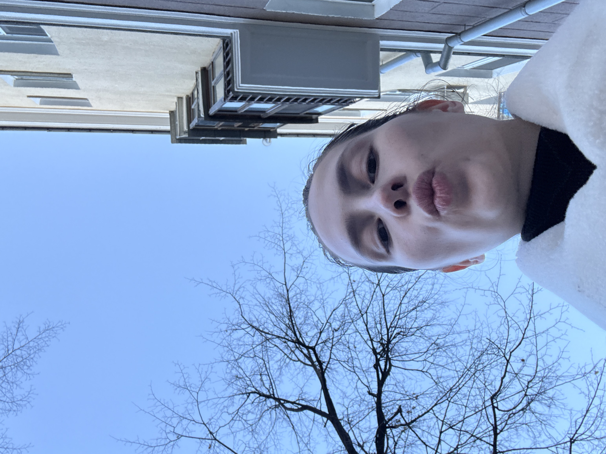 A selfie taken outdoors, likely in Yekaterinburg, Russia. A young woman with dark hair pulled back makes a pursed-lip face. The sky is light blue with bare branches of trees in the lower half of the frame. Above the woman is part of a building with light-colored walls and gray architectural details, including a balcony. She wears a dark shirt and a white coat or sweater.