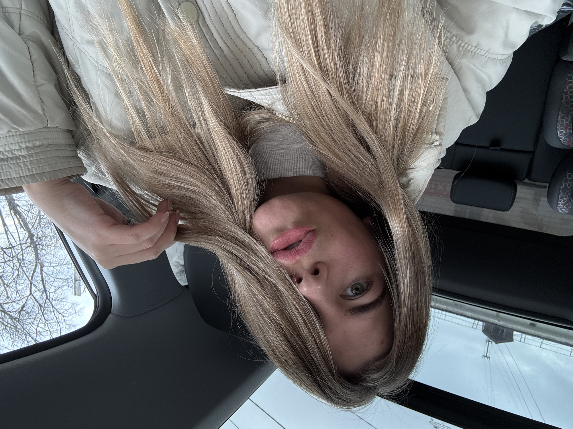 A young woman with long blonde hair is taking a selfie from inside a car. The photo is taken at an angle, making it appear upside down. The woman is wearing a light-colored jacket and a gray sweater underneath. Her face is visible, revealing light skin, pink lips, and green eyes. Her hand is visible in the top left corner of the frame, holding a strand of her hair. The background shows the interior of a car, including the back seats and the ceiling. Outside the car, bare trees and a cloudy sky are visible. A building with power lines can be seen in the reflection of a window. The photo appears to be taken in Chaikovsky, Russia.