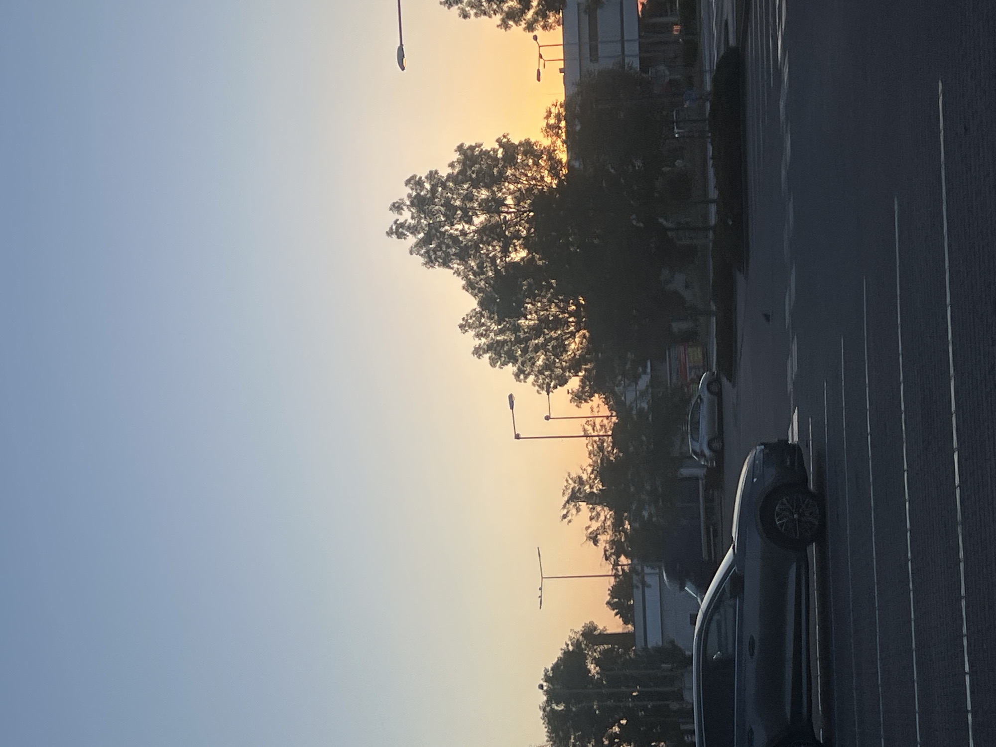 The image depicts a parking lot at dusk, likely in Trnava, Slovakia, given the metadata. The sky transitions from a clear blue at the top to a warm orange glow near the horizon, indicating sunset. Silhouetted against the sunset are several trees, streetlights, and buildings. In the foreground, a dark-colored SUV is parked in a marked parking space. Another car, a silver hatchback, is visible further in the background, also in a parking space. The parking lot is paved and marked with white lines. The overall scene suggests a common urban or suburban environment during the evening hours.