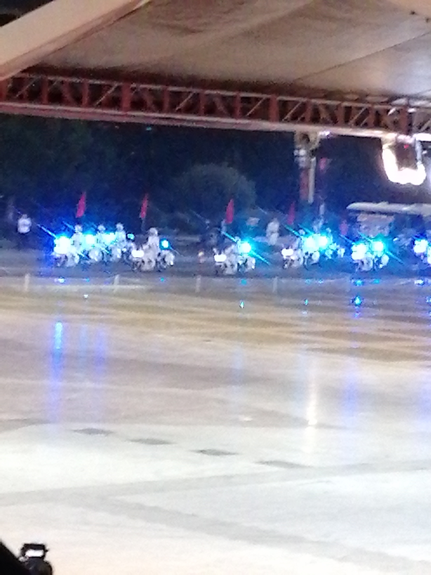 The image shows a nighttime outdoor event, possibly a parade or performance, in Da Lat, Vietnam. A group of people on motorcycles, dressed in what appears to be white uniforms, are illuminated by bright blue lights. The motorcycles themselves are also adorned with blue lights, creating a striking visual effect. Red flags are visible in the background, suggesting a festive or ceremonial occasion. The ground is a smooth, reflective surface, perhaps a stage or a wet pavement, which further enhances the lighting effects. The overall scene is dynamic and visually engaging, with the bright lights cutting through the darkness. There is no visible text in the image. The weather appears to be clear, allowing for an outdoor event. The presence of the motorcycles and uniformed riders suggests a synchronized performance or a display by a special unit.