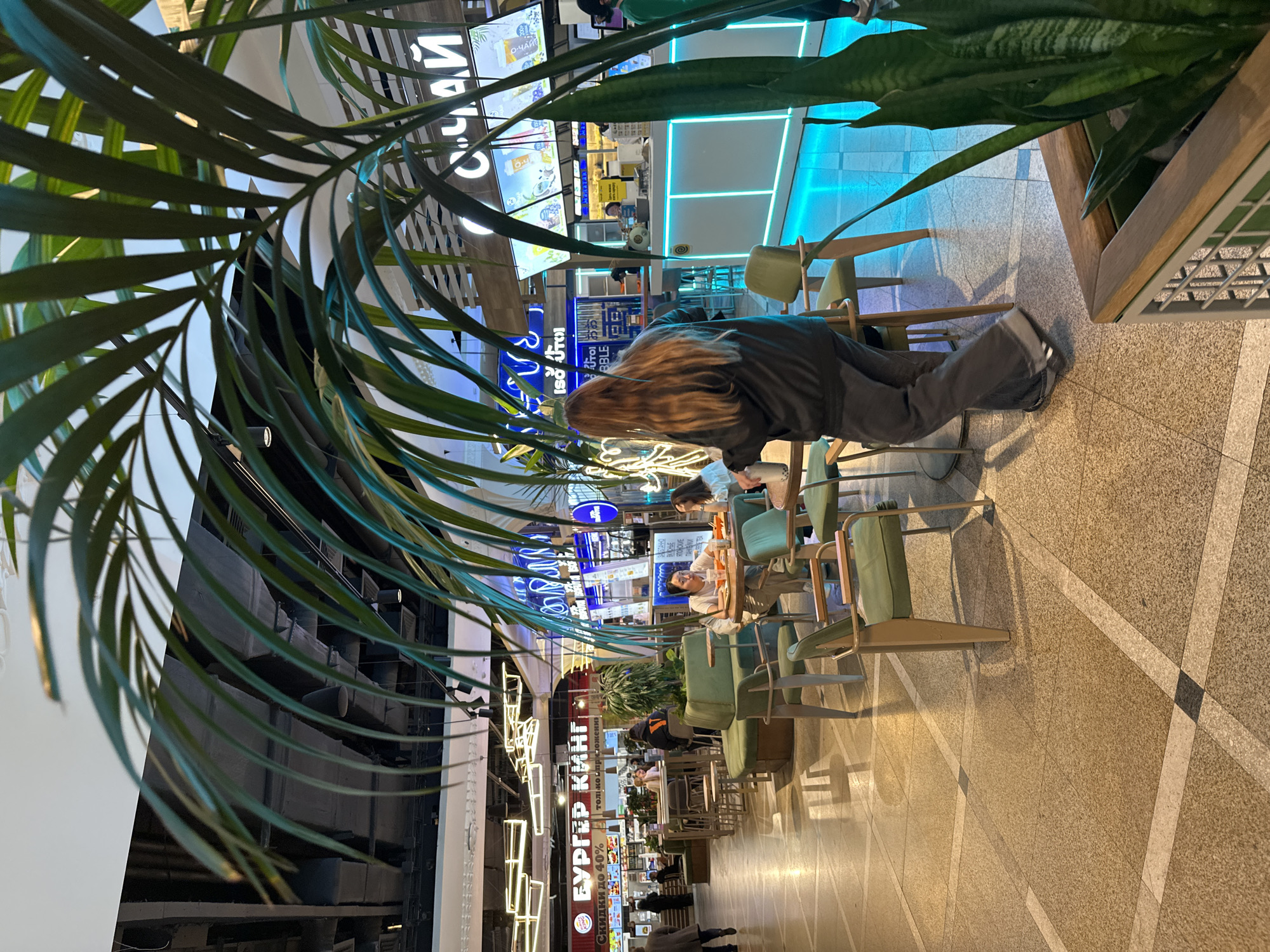 The image captures an indoor scene, likely a food court or mall corridor, viewed from a slightly elevated perspective. In the immediate foreground on the left, large, green palm-like leaves partially frame the shot.
The scene features a long walkway with a light-colored, geometrically patterned floor. Along both sides of this walkway, there are numerous small tables paired with light green chairs, forming casual dining areas. Several individuals are seated at these tables. One person with long, light brown hair, wearing a dark top and grey pants, is prominently featured in the mid-left, leaning forward over a table. Other people are visible further down the corridor, some appearing to be eating or looking at phones.
On the right side of the walkway, another large green plant in a wooden planter is visible. The background reveals various shopfronts and stalls, illuminated by bright, colorful neon signs, creating a vibrant atmosphere. Overhead, lighting fixtures cast a warm glow.
Visible text on the signs includes "БУРГЕР КИНГ" (Burger King) with an accompanying offer "Скидки до 40% только приложения" (Discounts up to 40% only through the app). Other signs show "О-ЧАЙ" (O-Chai), "СЧАЙ", "ЯПОНСКИЕ ПАНКЕЙКИ" (Japanese Pancakes), and partially visible "BBLE", likely referring to Bubble Tea. The presence of Russian text aligns with the given location context of Yekaterinburg, Russia. The overall impression is that of a bustling, well-lit commercial interior.