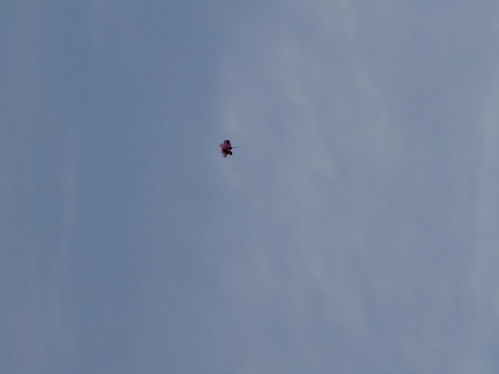 The image captures a cluster of pink or magenta balloons floating high in a vast, blue sky. The sky is largely clear, showing a consistent light blue hue, with faint traces of wispy white clouds barely visible. The balloons are positioned near the center of the frame, appearing to ascend or drift across the sky. There are no people, structures, or any other identifiable objects in the composition, emphasizing the emptiness and expanse of the sky. The time of day is clearly daytime, indicated by the bright and well-lit conditions. While the provided context specifies the location as Moscow, Russia, there are no visual cues within the image itself to confirm this. No text is visible.