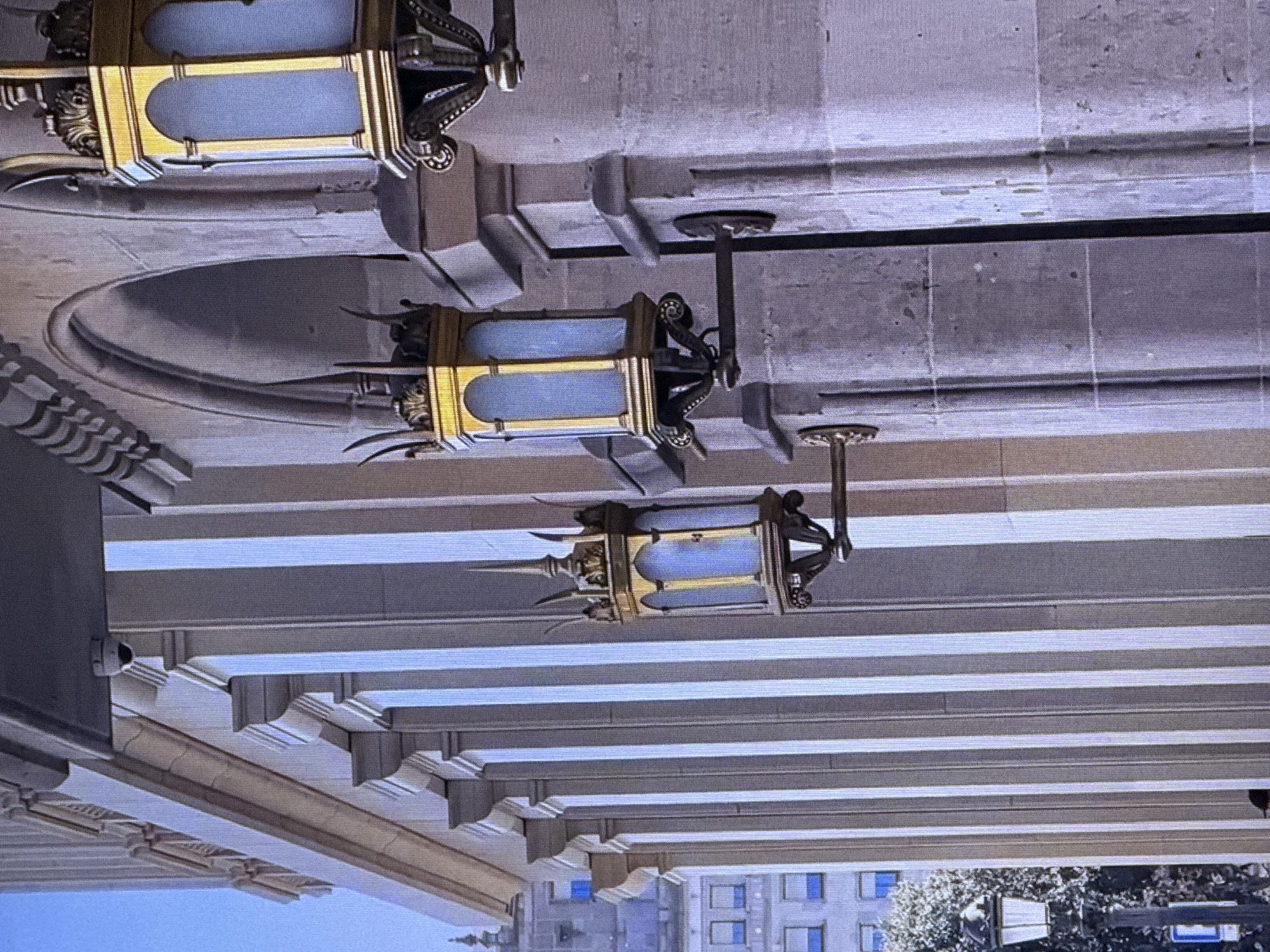 This media file depicts an outdoor scene with architectural elements and decorative lighting fixtures. The image is oriented such that the main subjects are upside down. The primary objects are ornate, gold-colored lanterns with blue glass panels, suspended from the ceiling of a building's overhang. These lanterns are intricate, featuring decorative scrollwork and pointed finials.
The setting appears to be a grand, possibly historic, building with a stone facade and a series of evenly spaced, slatted overhangs or beams. There are also decorative moldings and architectural details visible along the walls and ceiling. In the background, through an opening formed by the architectural structure, we can see a glimpse of a city skyline. Buildings with visible windows, some blue, are present, along with trees and a street sign.
The time of day is unclear due to the lighting, but it appears to be daytime. There are no people or activities explicitly shown. The overall impression is one of architectural grandeur and decorative ornamentation. The context provided indicates the location is Dubai, United Arab Emirates. There is no visible text in the image.