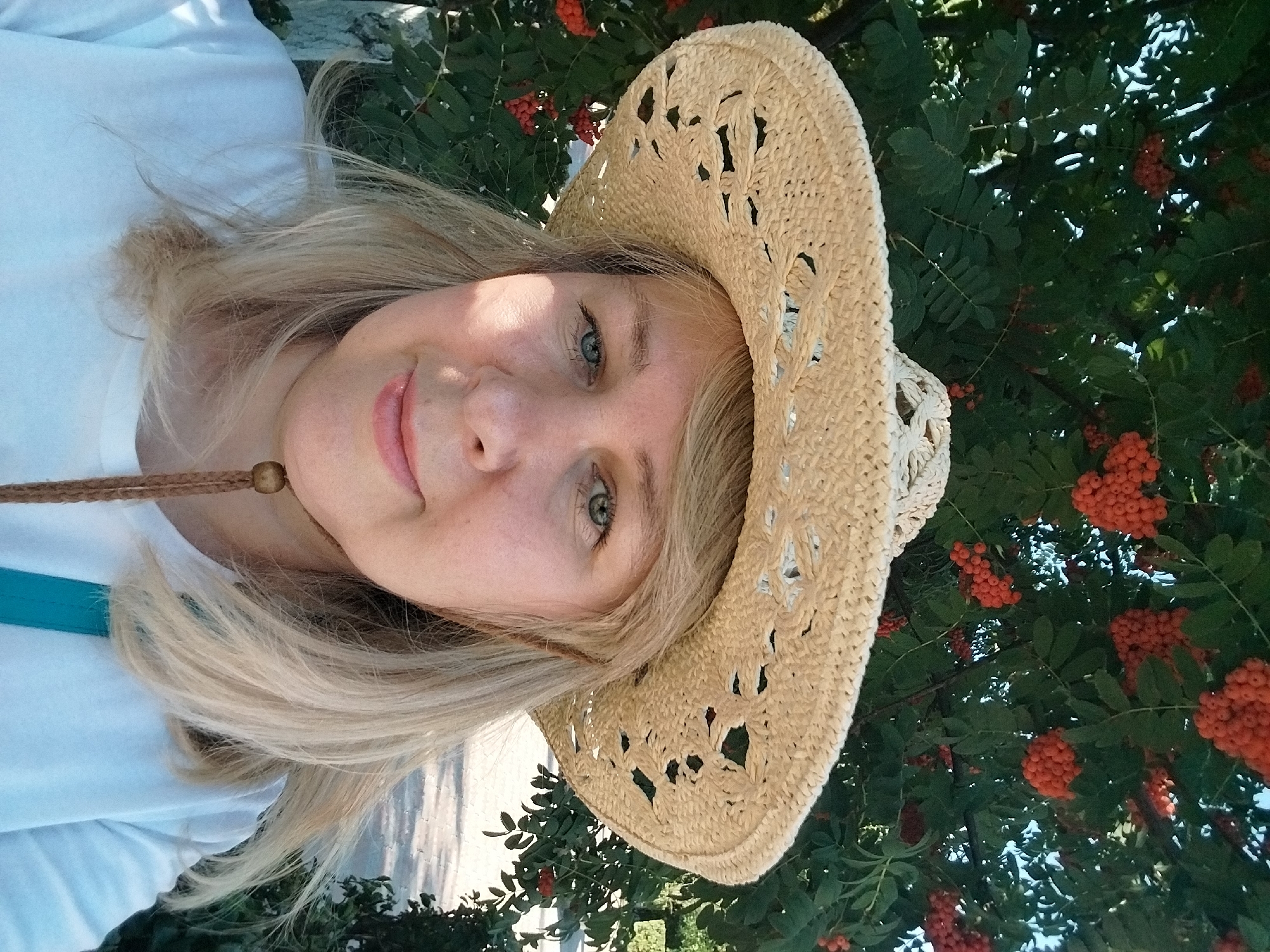 A medium shot captures a blonde woman wearing a wide-brimmed straw hat and a white t-shirt, posing for a selfie outdoors. Her face is tilted slightly to the left, and she is looking at the camera with light blue eyes. Behind her, a tree laden with clusters of bright orange berries and green leaves forms a natural backdrop. The scene appears to be in a park or garden setting, likely during daylight hours given the bright lighting. The woman has a light complexion and subtle makeup, with her blonde hair visible beneath the hat. A brown cord with a wooden bead hangs down from her hat, crossing her chest. The background is slightly blurred, suggesting a shallow depth of field, which focuses attention on the woman. The overall impression is of a pleasant, casual outdoor moment. The location in Orenburg, Russia, would suggest a temperate climate, and the presence of rowan berries indicates a late summer or early autumn setting.