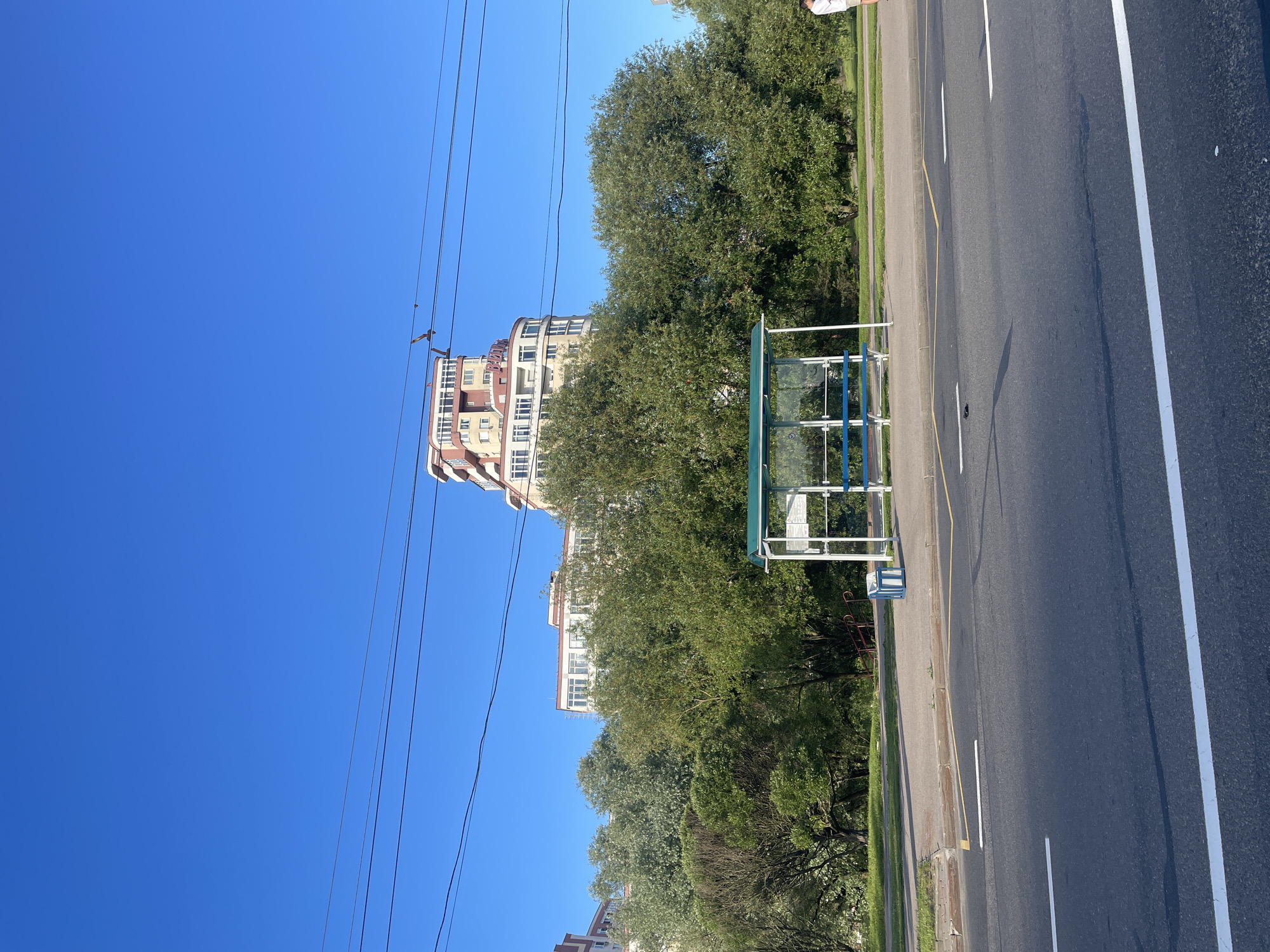 This media file depicts a bright, sunny day in Minsk, Belarus, likely during late morning or early afternoon given the clear blue sky and strong shadows. The scene is a roadside, showing a paved road with white lane markings and a yellow line along the shoulder. To the left of the road is a bus stop shelter with a green roof and glass walls, and a blue bench inside. Adjacent to the bus stop is a sidewalk and a grassy verge lined with lush green trees. Behind the trees, a tall, modern apartment building with a distinctive facade featuring red and cream-colored sections and many windows can be seen. Power lines are strung across the sky, adding to the urban feel. The overall atmosphere is one of everyday life in a city, with the clear weather suggesting a pleasant day. There is no visible text.