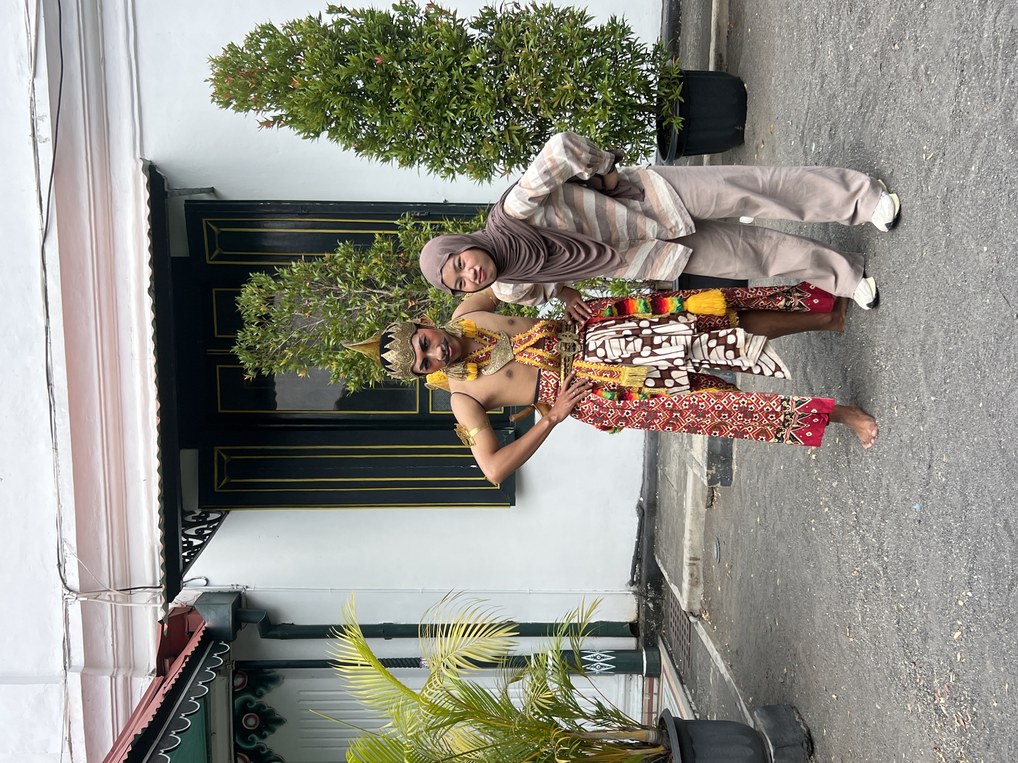 Two people are posing for a photo in front of a white building with black and gold-trimmed windows. The person on the left is a man dressed in traditional Javanese attire, including a headdress, elaborate garment, and patterned pants. He is barefoot. The person on the right is a woman wearing a hijab and a striped shirt with loose-fitting pants. They are both looking towards the camera, with their hands on their hips. To the left of the man is a potted palm plant, and to the right of the woman is a potted evergreen tree. The ground is paved with asphalt. The building appears to be historical or cultural in nature, possibly located in a prominent area of Yogyakarta, Indonesia, given the attire and architectural style.