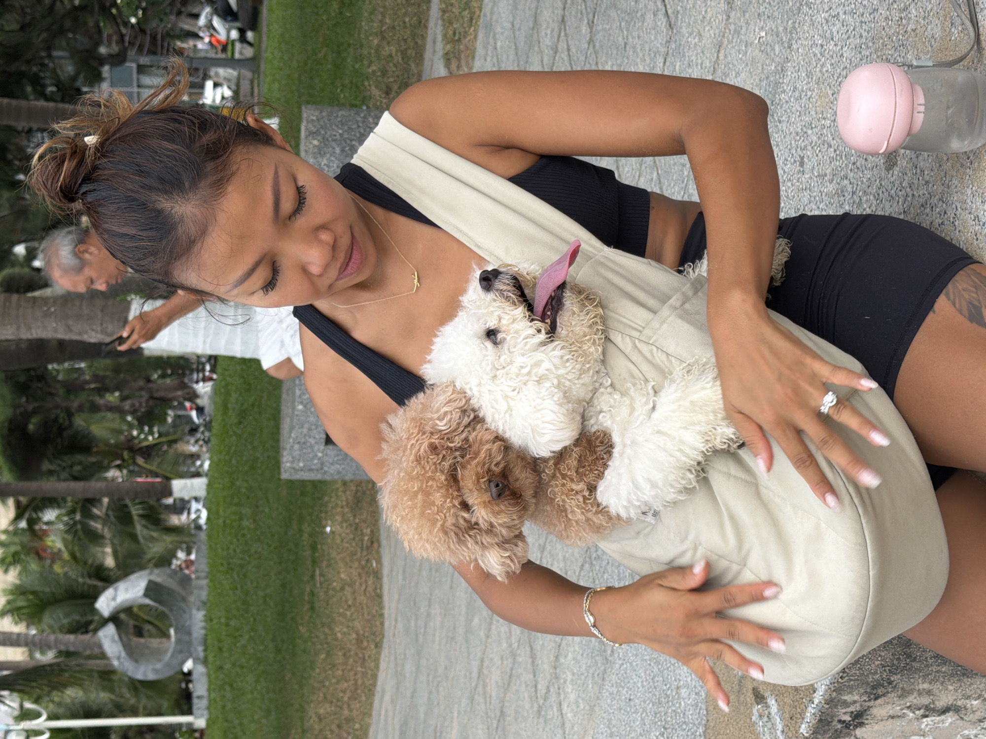 A woman is sitting outdoors in Nha Trang, Vietnam, holding two small dogs in a fabric carrier. The woman has her hair in a bun and is wearing a black tank top and black shorts. She has a bracelet on her left wrist and a ring on her left ring finger. One dog is brown and the other is white. In the background, there are palm trees, grass, and a sculpture. A man is visible in the background on the left, looking at his phone. A pink portable water dispenser is on the ground to the right of the woman. The setting appears to be a park or public area.