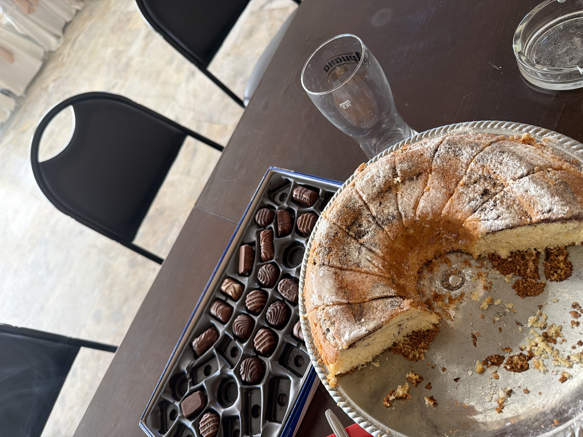 A top-down view shows a partly eaten ring cake on a silver platter alongside a box of chocolates and a beer glass on a dark brown table. An ashtray sits near the cake. Two black chairs are visible on the left. The background suggests an indoor space with a light-colored floor. There is no specific visual information to definitively place the scene in Varna, Bulgaria.