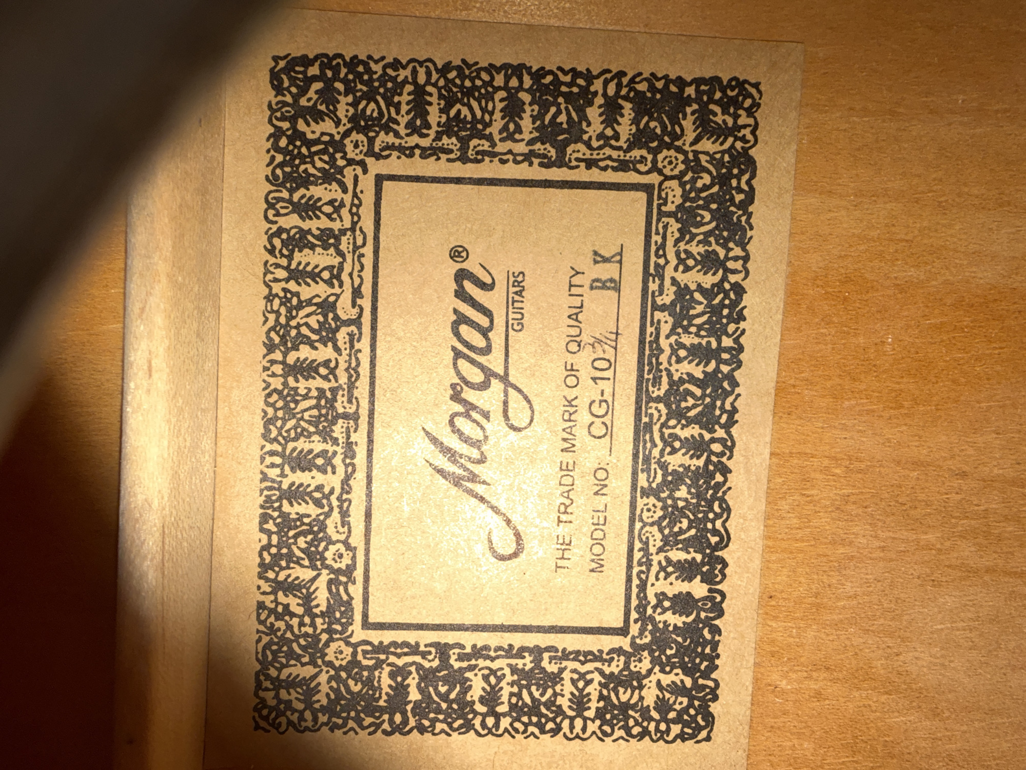 The image shows a close-up of a label inside a wooden instrument case. The label is tan with a decorative black border. In the center of the label, the word "Morgan" is printed in a stylized script, followed by the word "GUITARS" in smaller block letters. Below this, it reads "THE TRADE MARK OF QUALITY" and "MODEL NO: CG-103/4 BK". The label is affixed to a light brown wooden surface. The lighting is warm, casting shadows from the edges of the label. There is no visible connection to Oslo, Norway.
