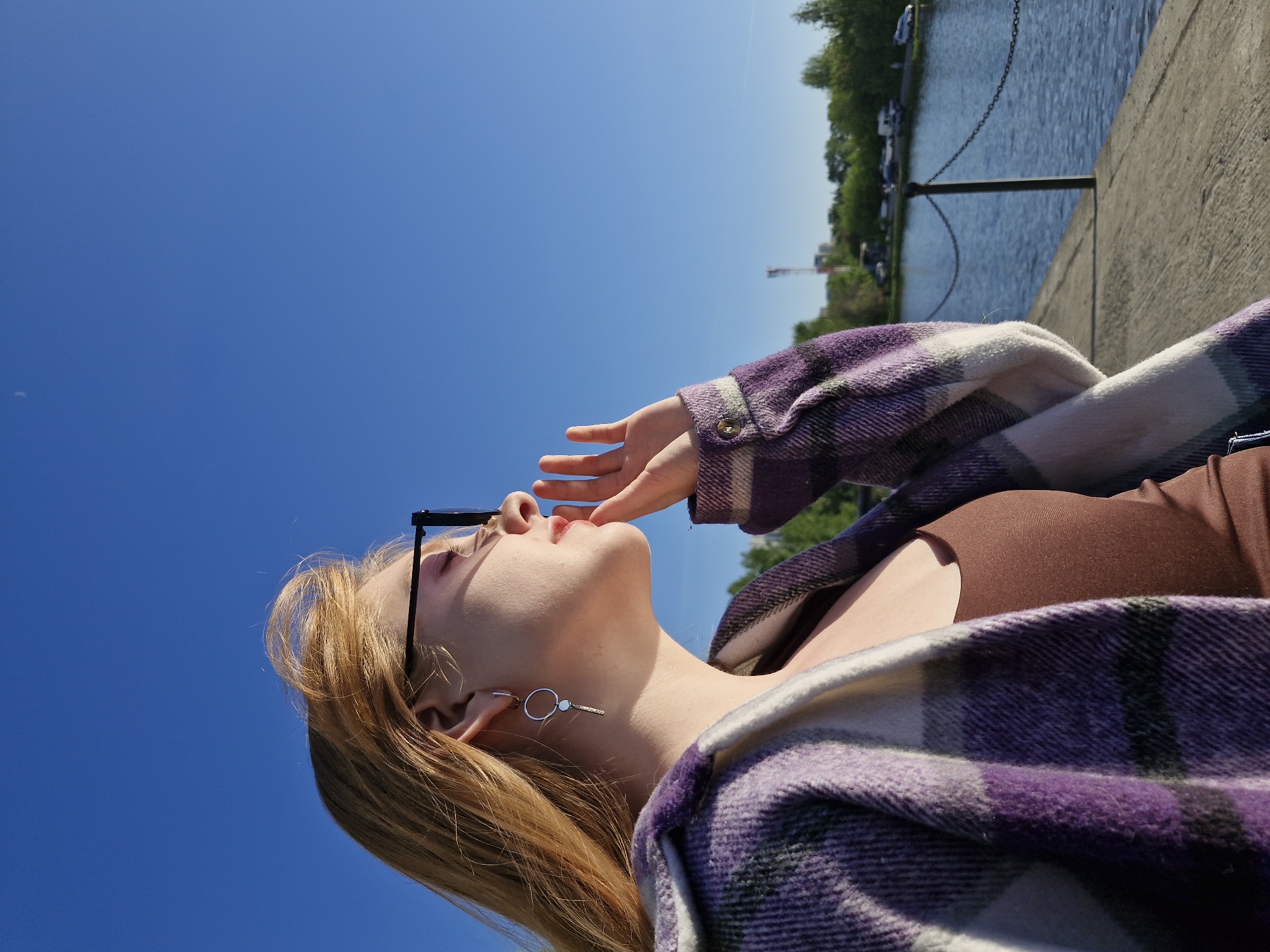 A fair-skinned woman with reddish-blonde hair is reclining outdoors, looking upwards. She is wearing eyeglasses and a purple and white plaid flannel shirt over a brown top. Her hand is raised near her face. A silver earring is visible in her left ear.
The background consists of a clear blue sky, a concrete embankment, and water, with trees and buildings visible in the distance.
The location is likely the city of Yekaterinburg, Russia, as it contains a body of water and green area and is a common location for pictures.
